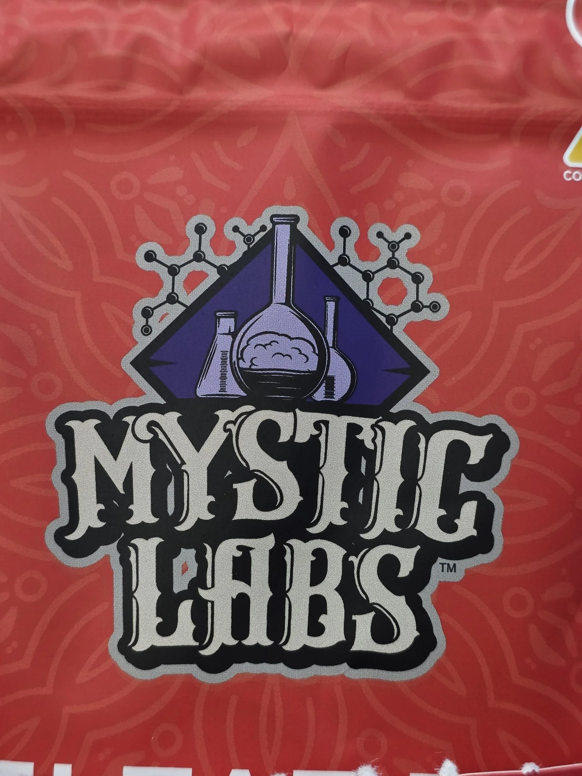 Close-up of a red package with a science-themed sticker featuring a laboratory flask and molecular structure, and some black and white text that says mystic-labs.