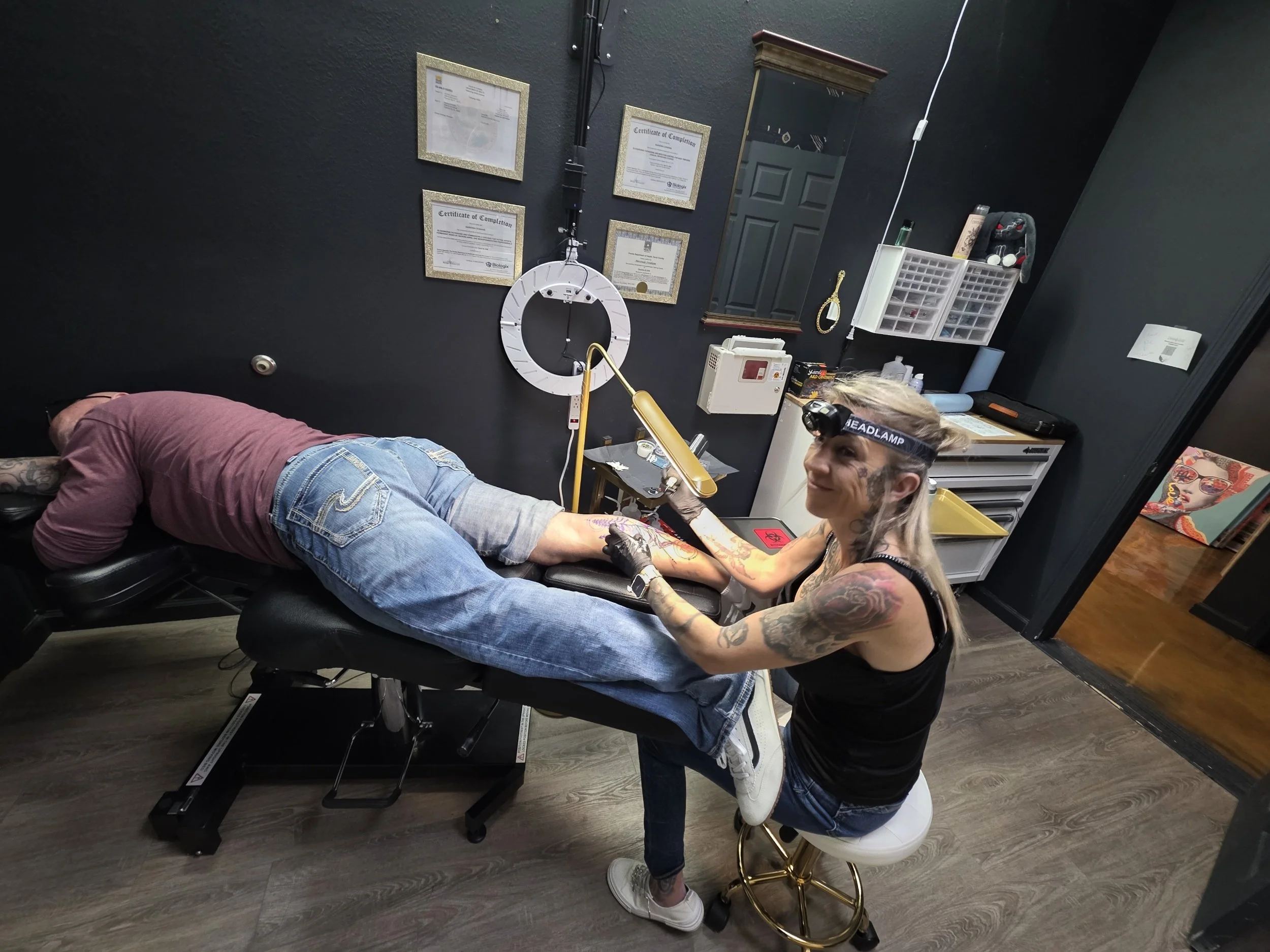 Tattooed woman with headlamp tattooing a man's leg during a tattooing session at Divinely Ink'd tattoo studio.
