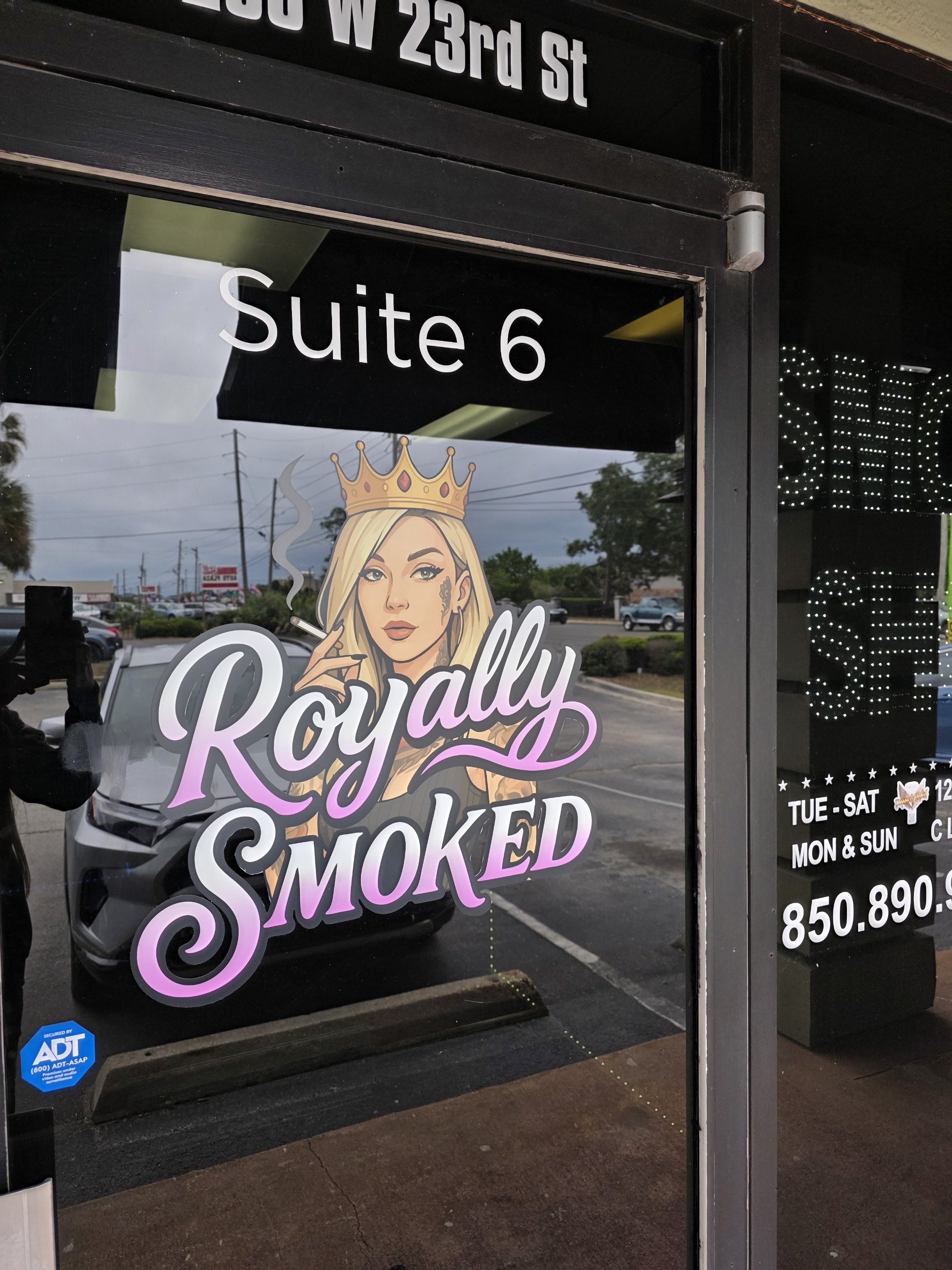Store window with a sign that says 'Suite 6' and a graphic of a woman with blonde hair wearing a crown, holding a cigarette, with the words 'Royally Smoked' in stylized font. The background shows a parking lot and cloudy sky.