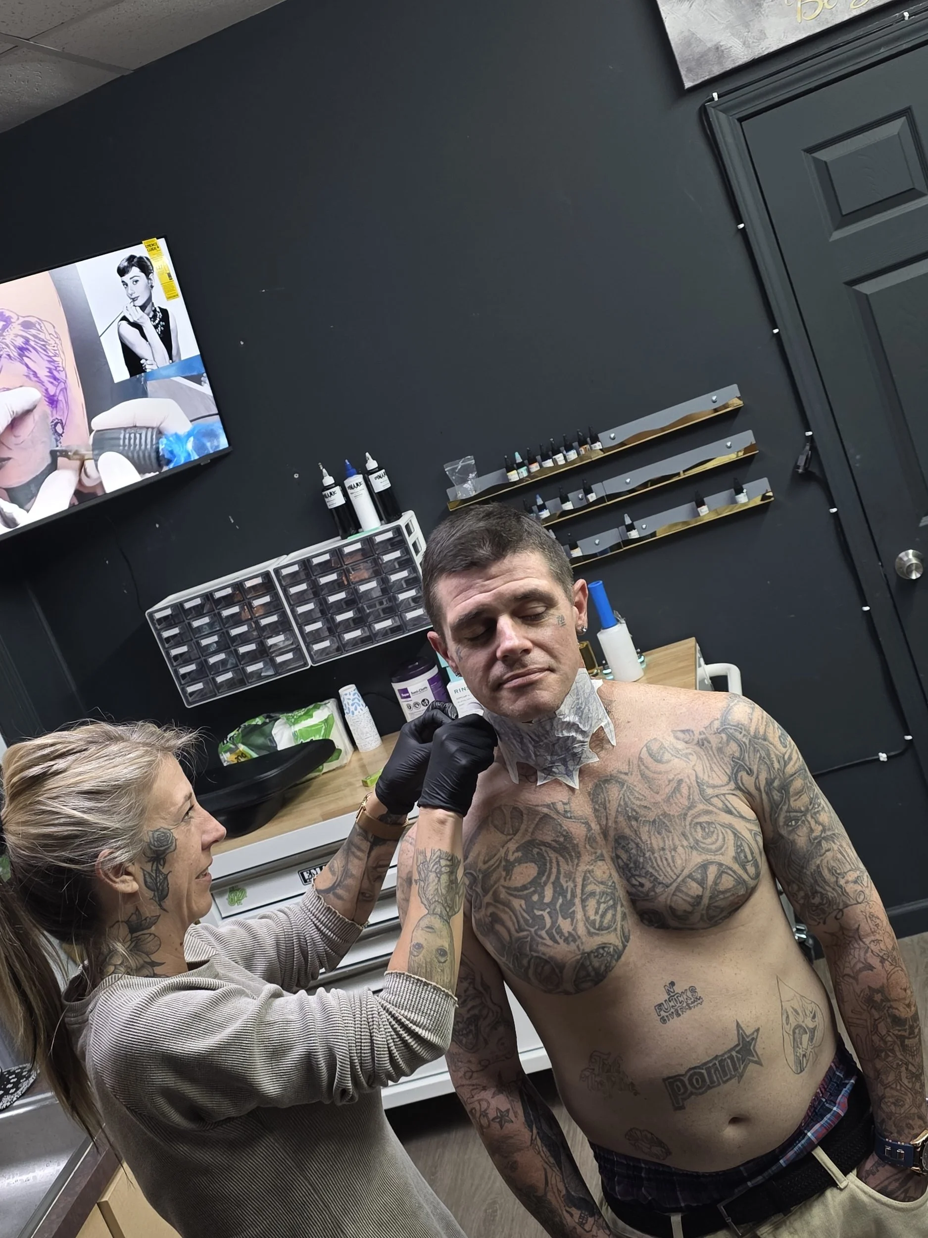 A man with tattoos on his chest and arms sitting shirtless in a tattoo studio, getting a tattoo applied to his neck by a tattoo artist. The man appears relaxed, and the tattoo artist is wearing gloves. The studio has black walls, shelves with tattoo 