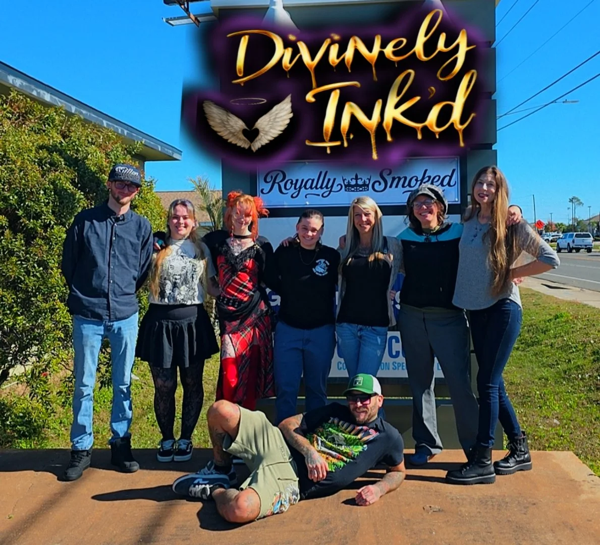 Group of eight people standing in front of a sign that reads "Divinely Ink'd" with angel wings logo, outdoors on a sunny day.