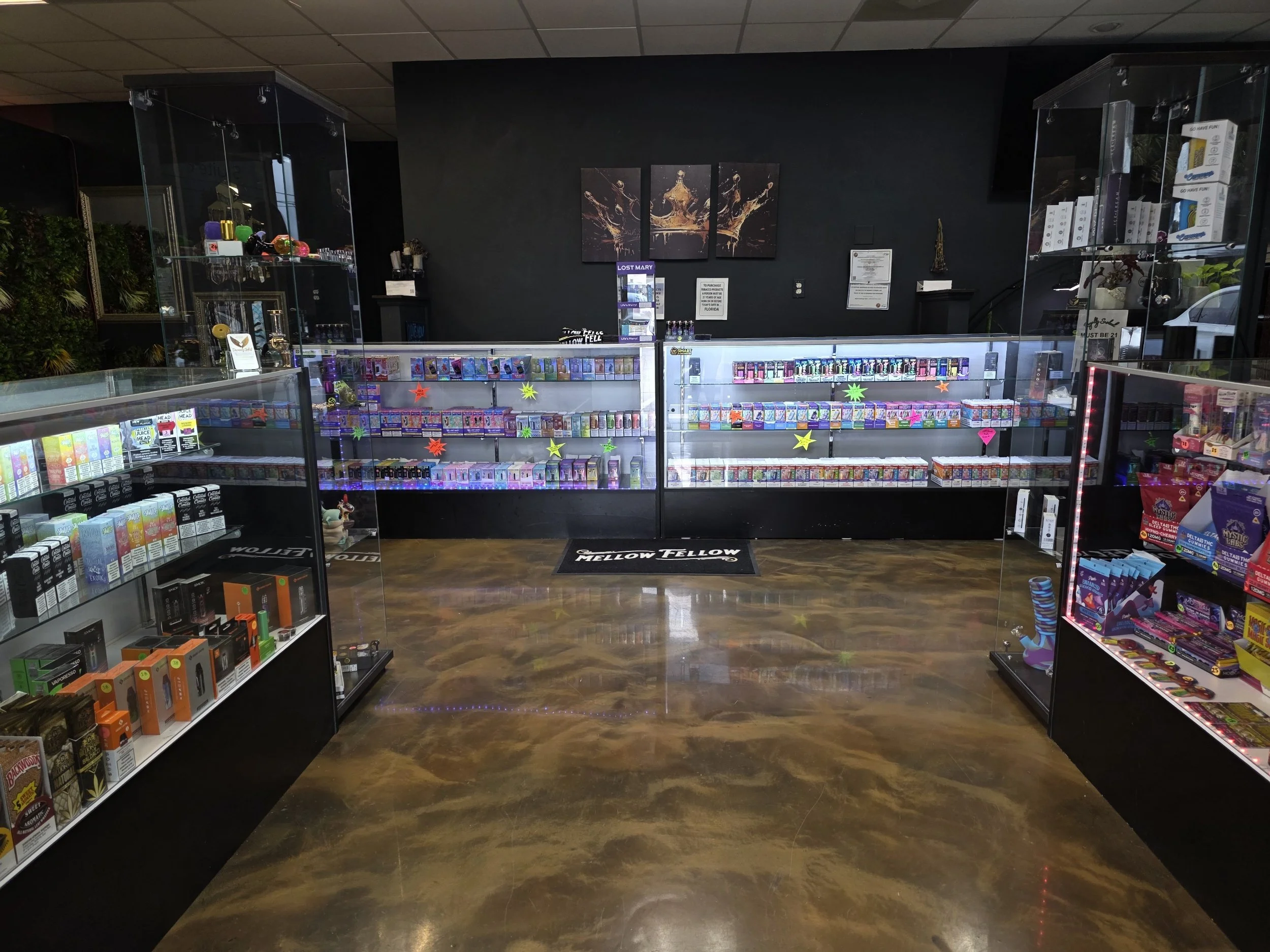 Inside a vape shop with glass display cases filled with vaping products and accessories. The background features dark walls with art and informational posters, and a wooden floor.