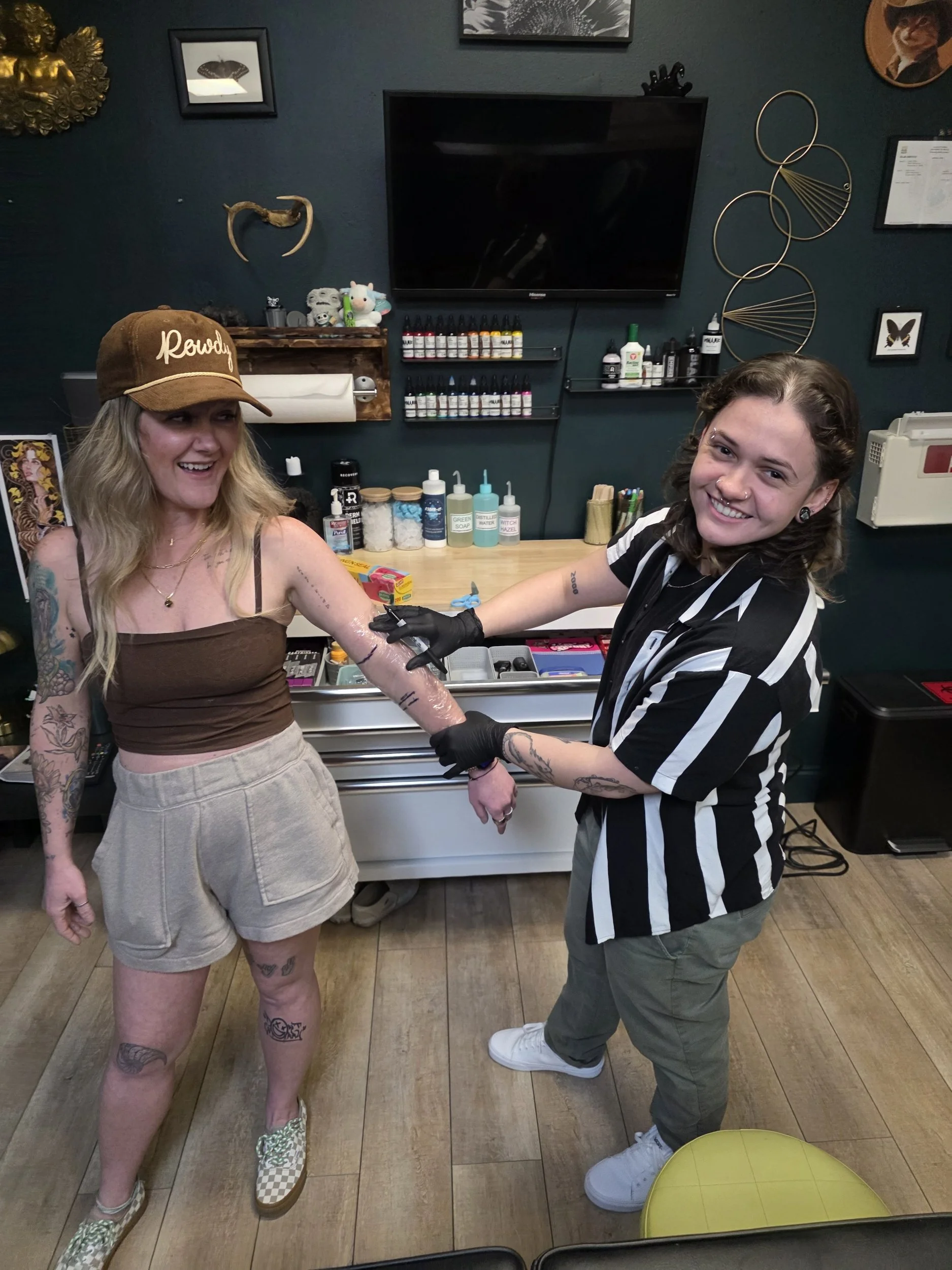 Two women are smiling at each other, one of whom is receiving a tattoo on her arm from the other. The woman receiving the tattoo has blonde hair, multiple tattoos on her arms and legs, and is wearing a brown tank top, beige shorts, and checkered slip
