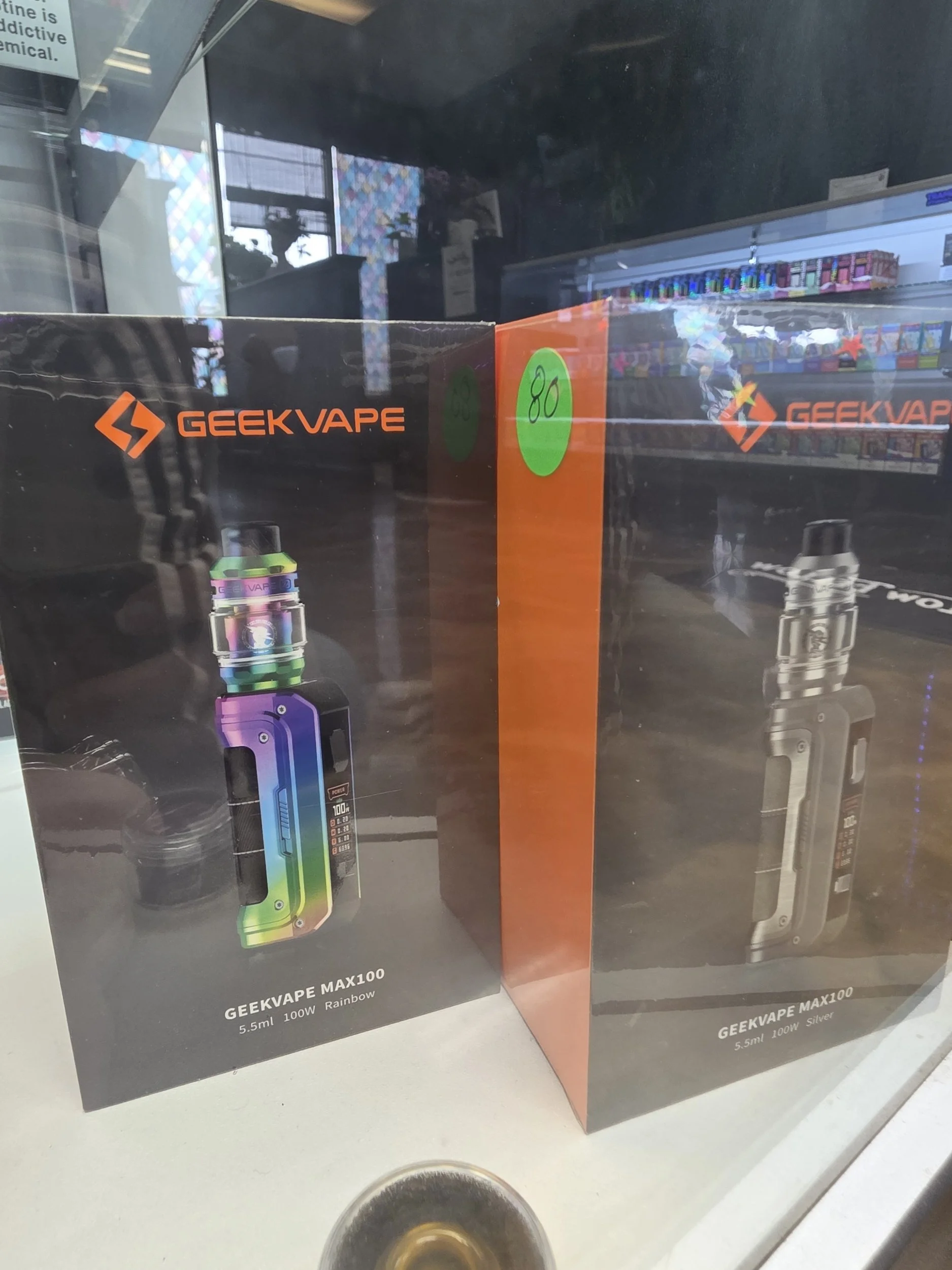 Boxes of GeekVape Max 100 rainbow and silver vape devices on display in a store window.