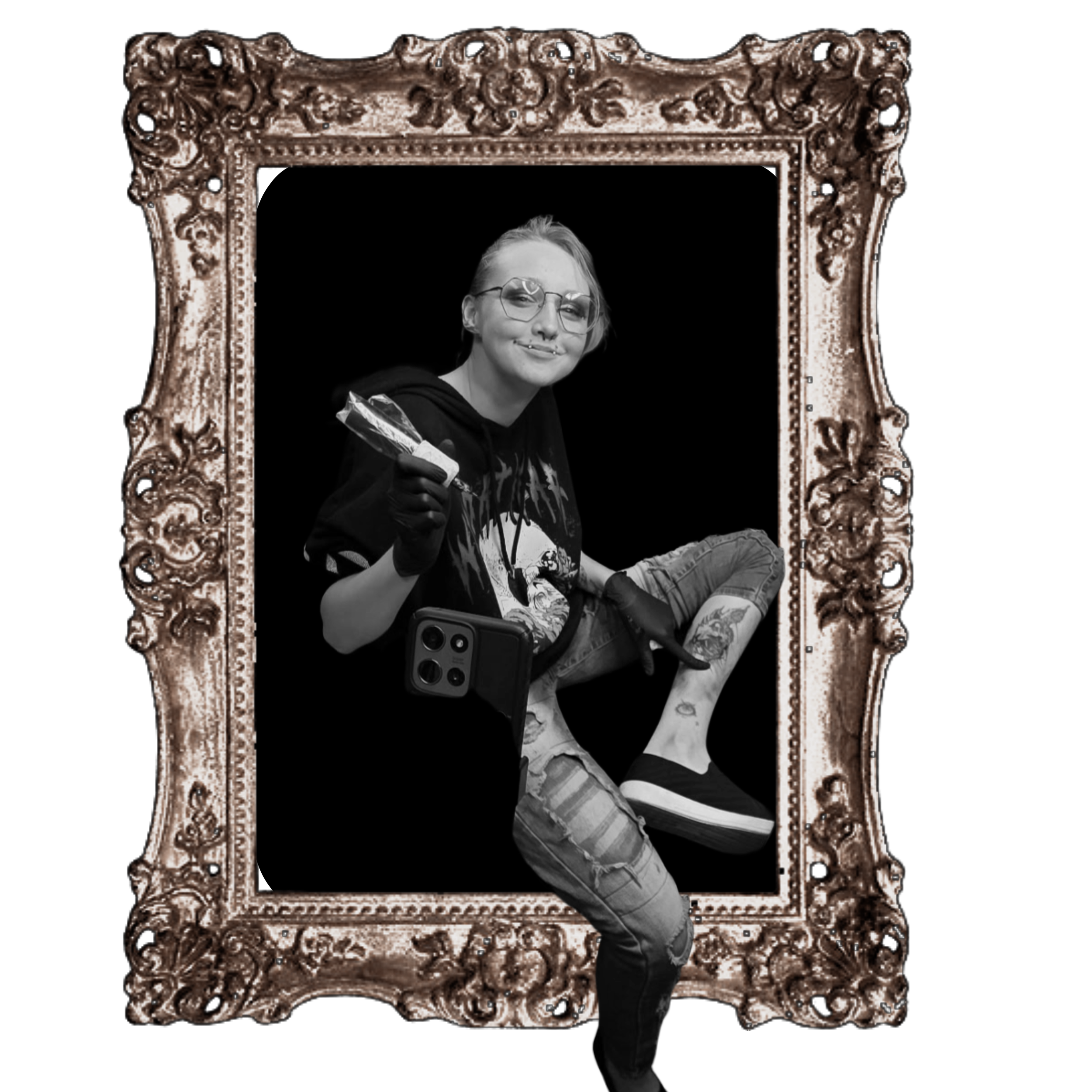 Kai with glasses, tattoos, and piercings posing inside an ornate, decorative picture frame, holding a tattoo machine and looking at the camera with a slight smile.