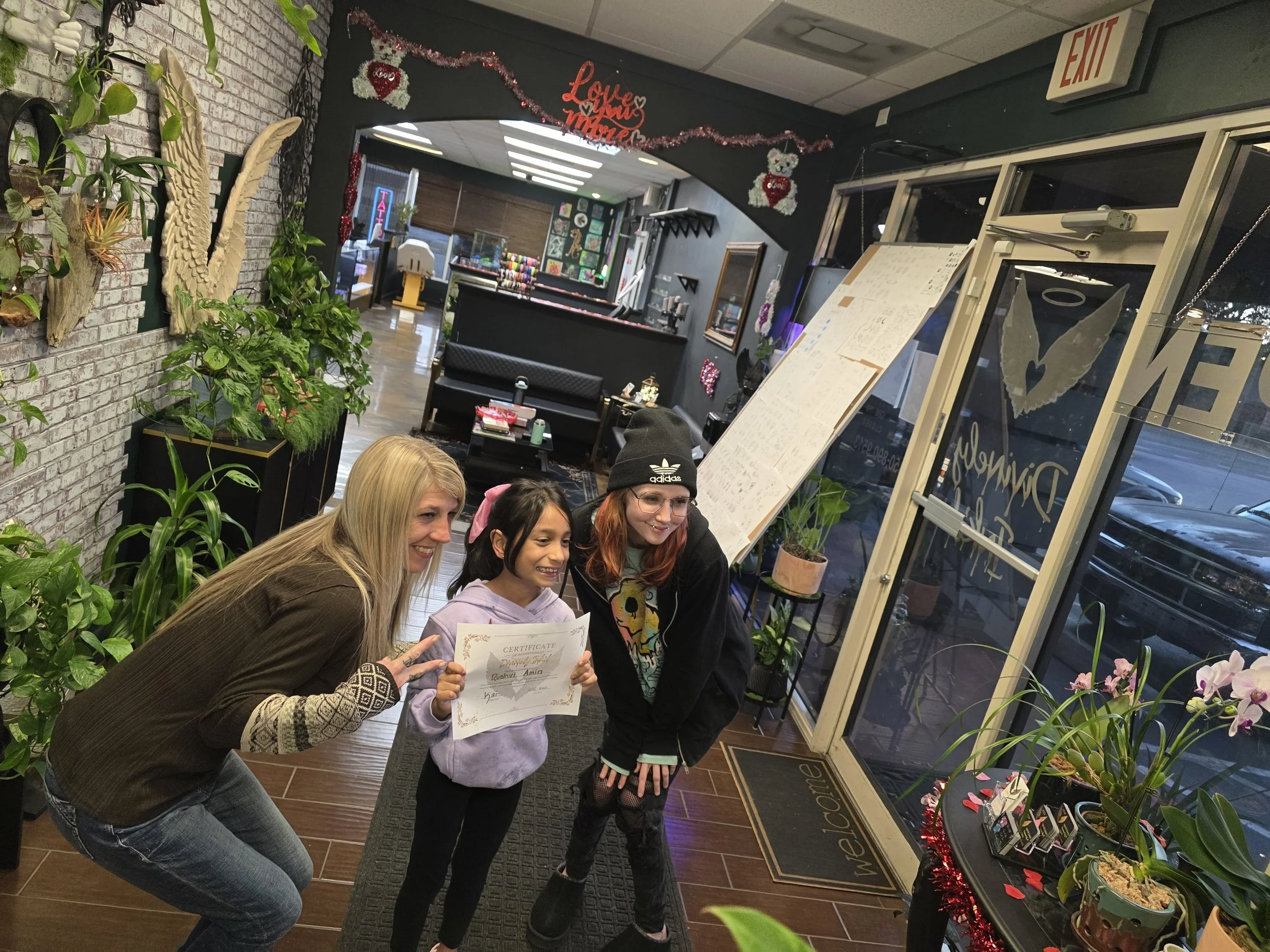 Three people are gathered in the lobby of a tattoo shop, smiling and posing for a photo. One person is holding a certificate, and there are plants and flowers around them. The background shows open spaces and wall decor.
