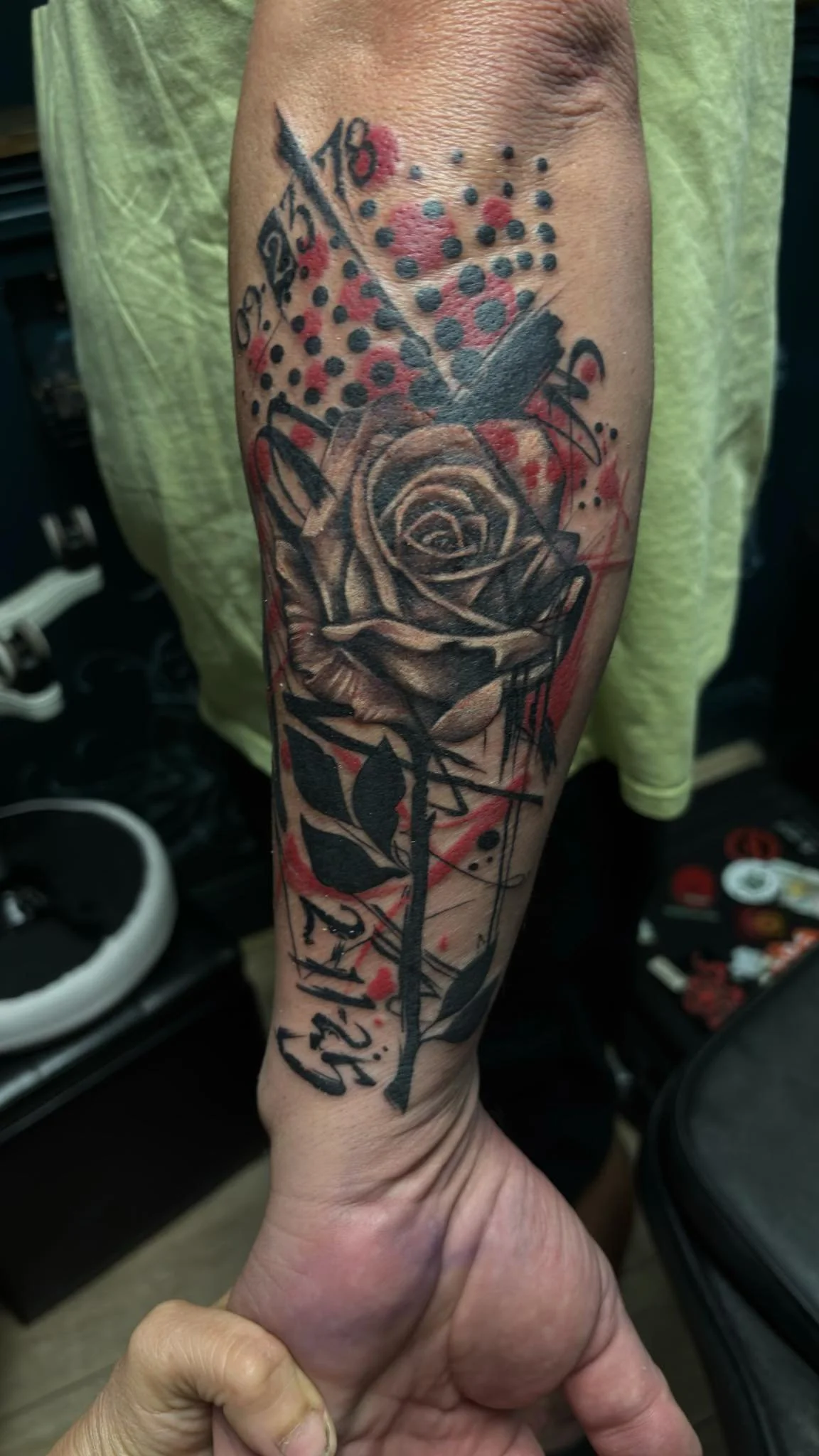A tattoo of a realistic black and gray rose with a dagger piercing through it on a person's forearm, surrounded by red and black abstract splashes and dots, with some black script at the bottom.