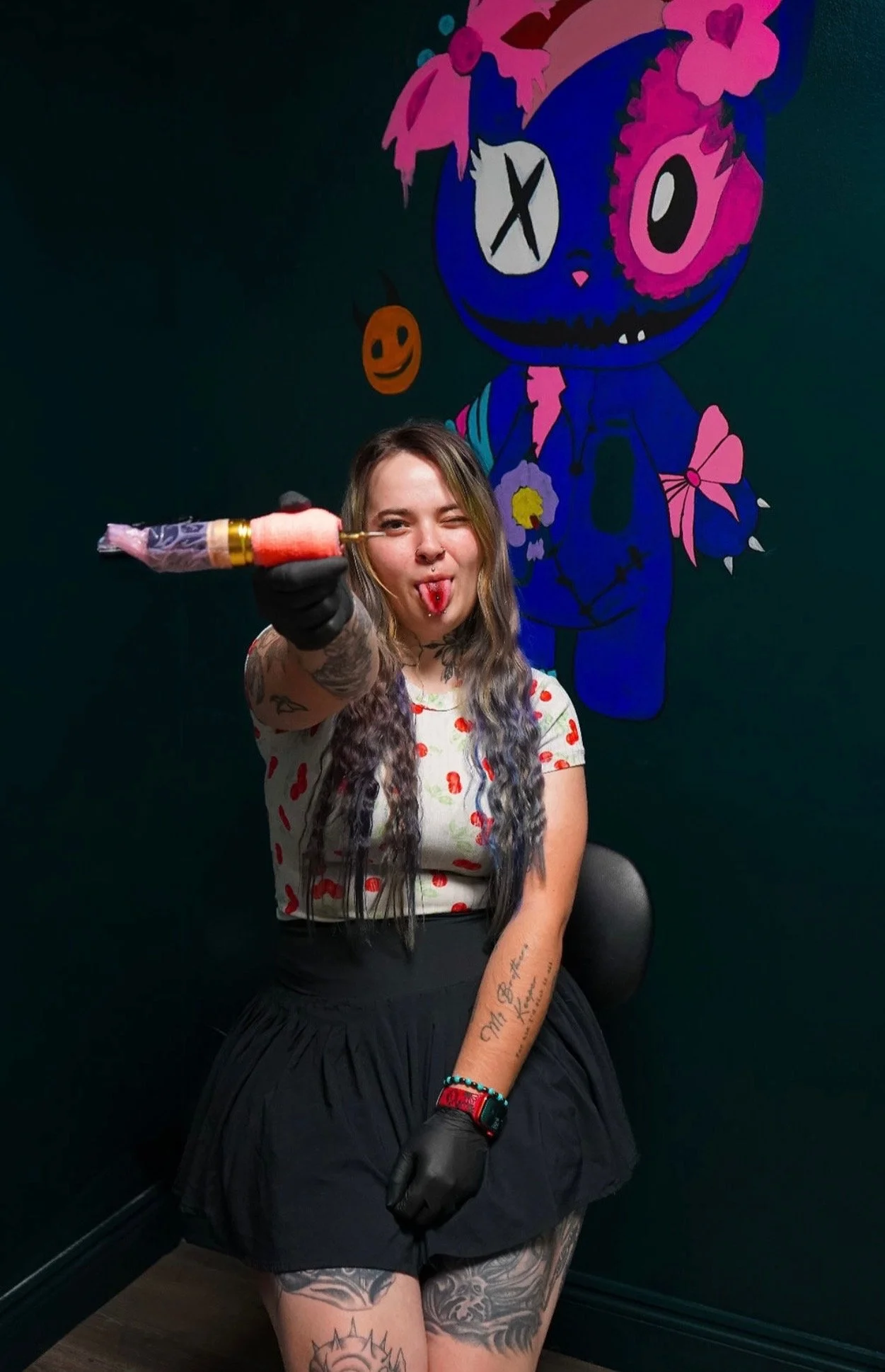 A young woman with tattoos and long, wavy hair, dressed in a white t-shirt with red heart patterns and a black skirt, is sitting on a chair. She is wearing a black glove on her right hand, holding a tattoo machine, and is winking with her tongue out.