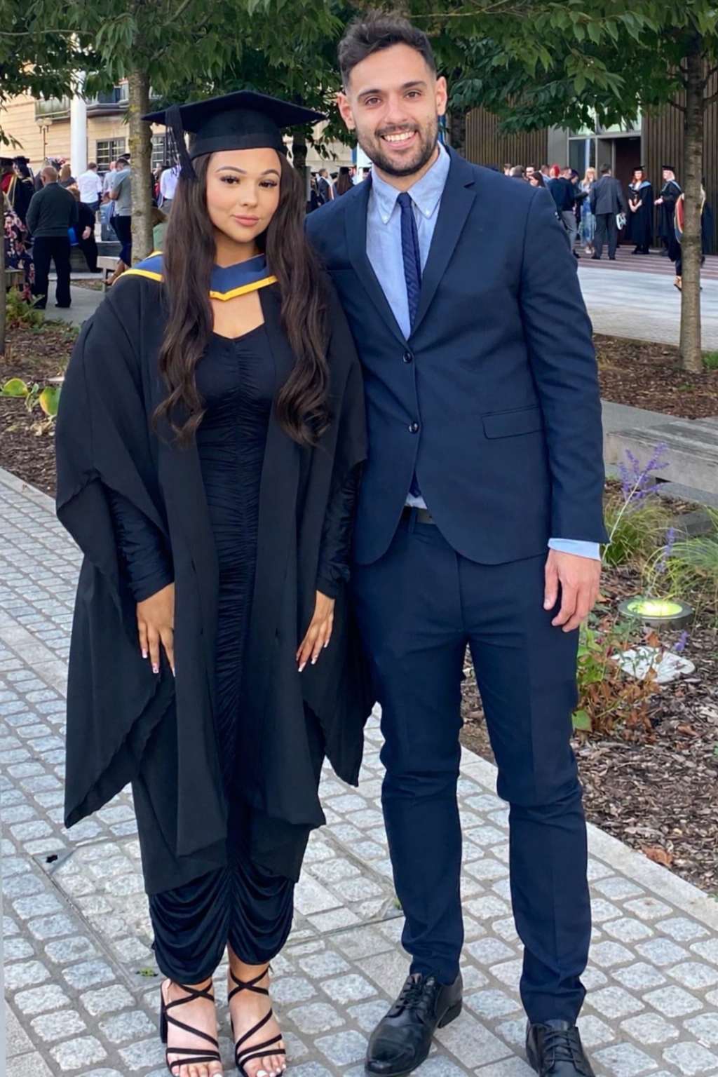A young woman in a black graduation gown and cap standing next to a young man in a suit, outdoors on a paved walkway with trees and other graduates in the background.