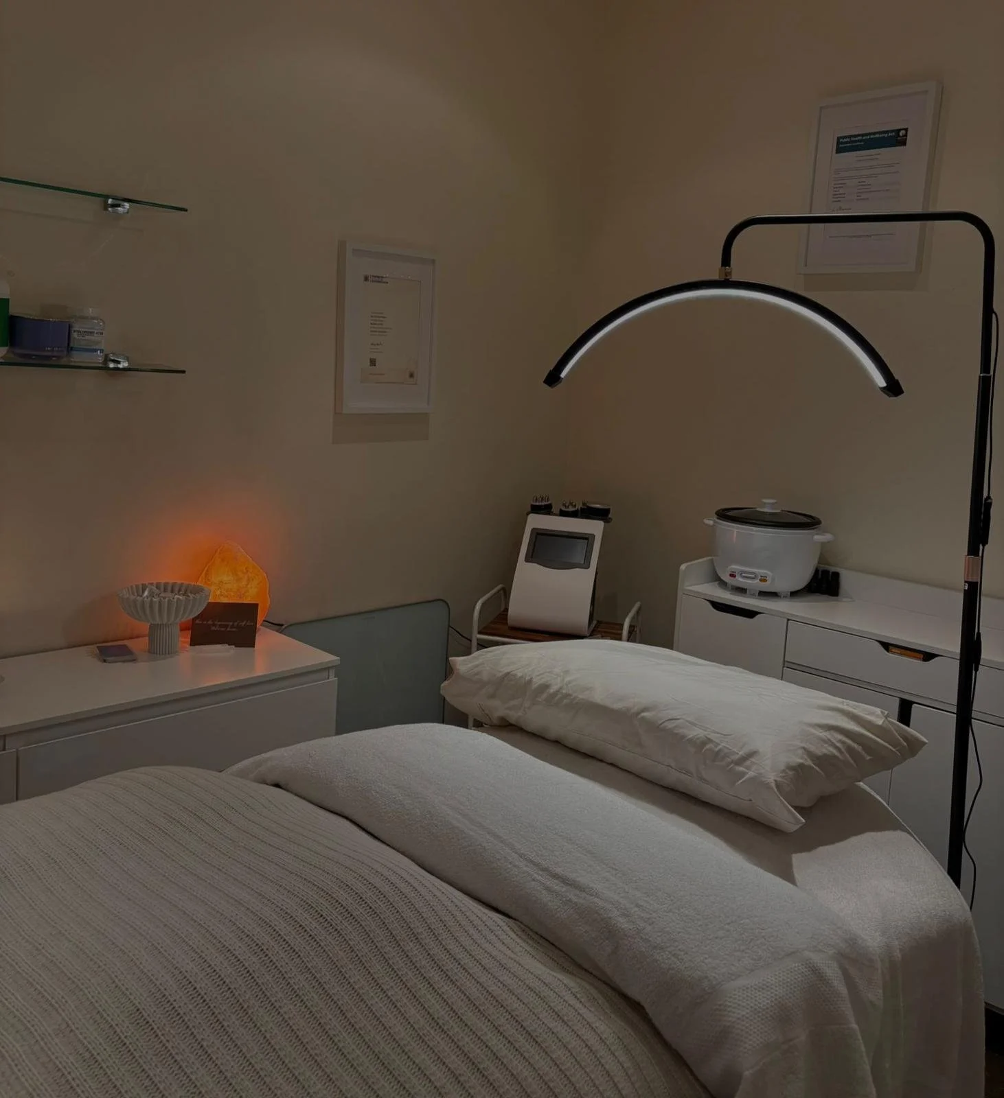 A hospital room with a bed, pillows, a side table with a Himalayan salt lamp, framed certificates on the walls, a lamp, a medical device, and a rice cooker on a cabinet.