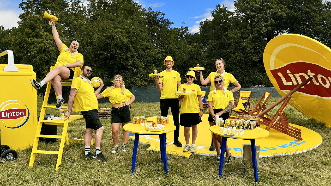 Group of people at a Lipton promotional event outdoors, wearing yellow shirts, with Lipton branded umbrellas, tables, and decorations, holding drinks and smiling.