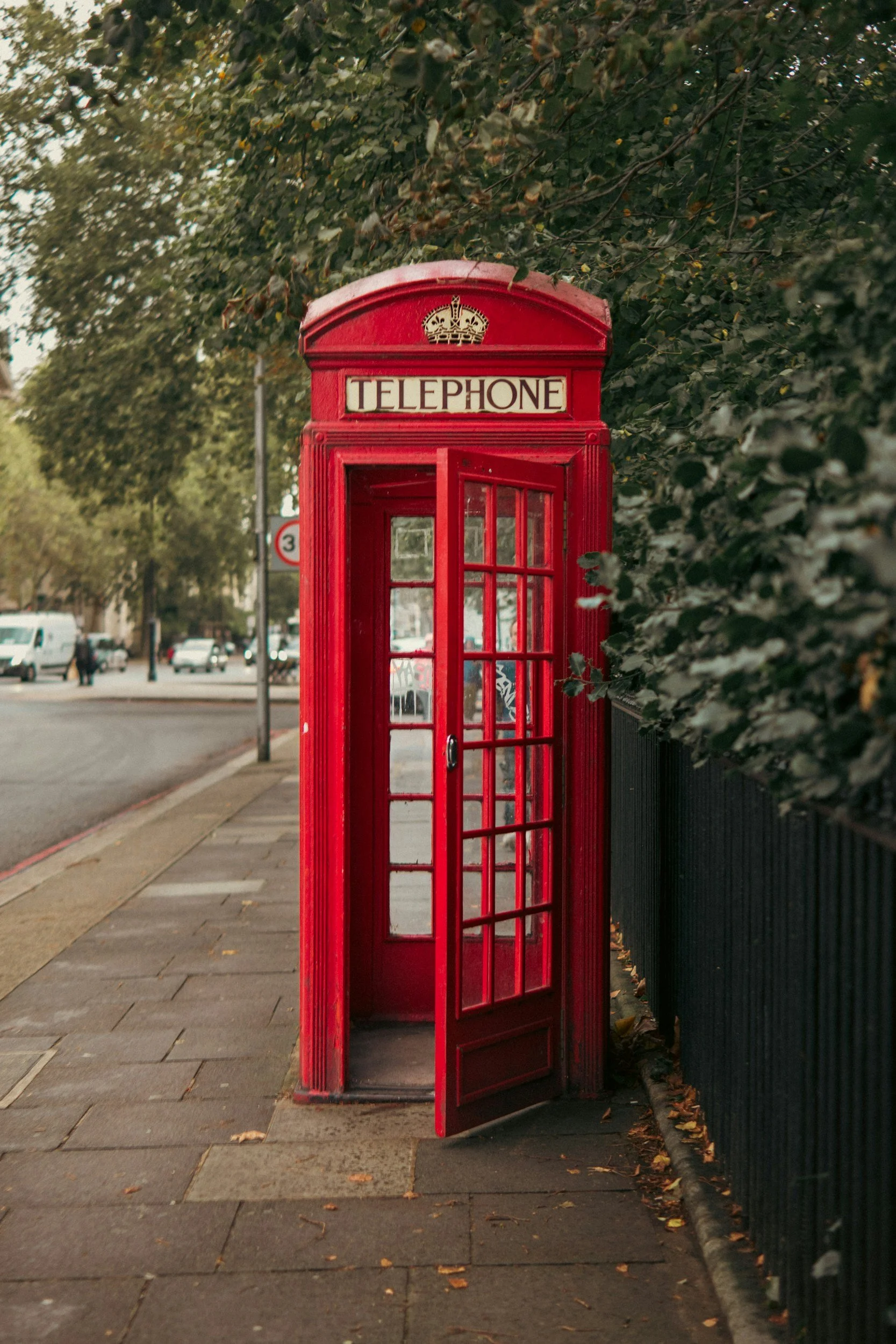 Red British telephone booth on sidewalk with trees and cars in the background
