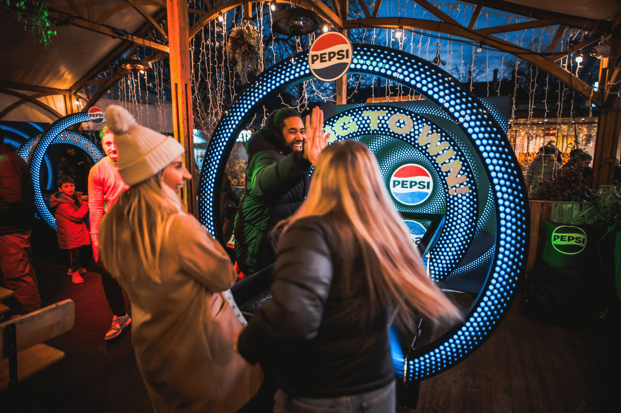 People smiling and giving high-fives in front of a illuminated Pepsi sign at an outdoor winter event with decorations and other guests in the background.
