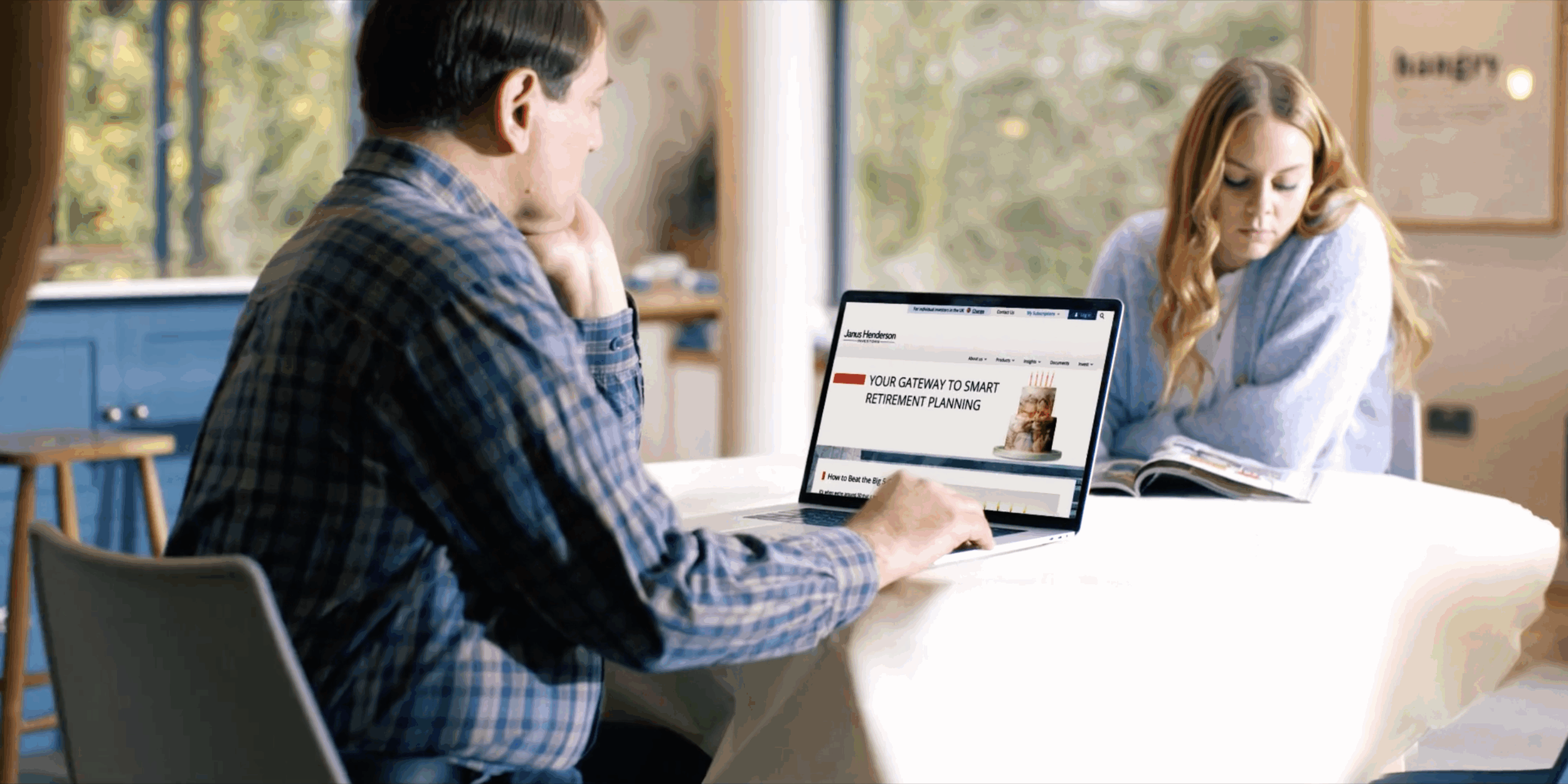 Two people sitting at a table, one man using a laptop with a retirement planning website open, and a woman looking at a magazine, in a well-lit room with large windows and trees outside.