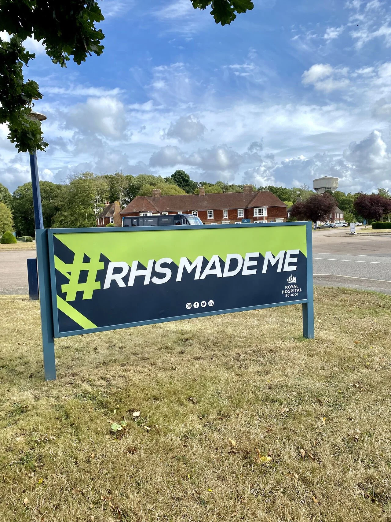 A large sign on a grassy area with text '#RHSMADE' and the Royal Hospital School logo, surrounded by trees and buildings in the background.