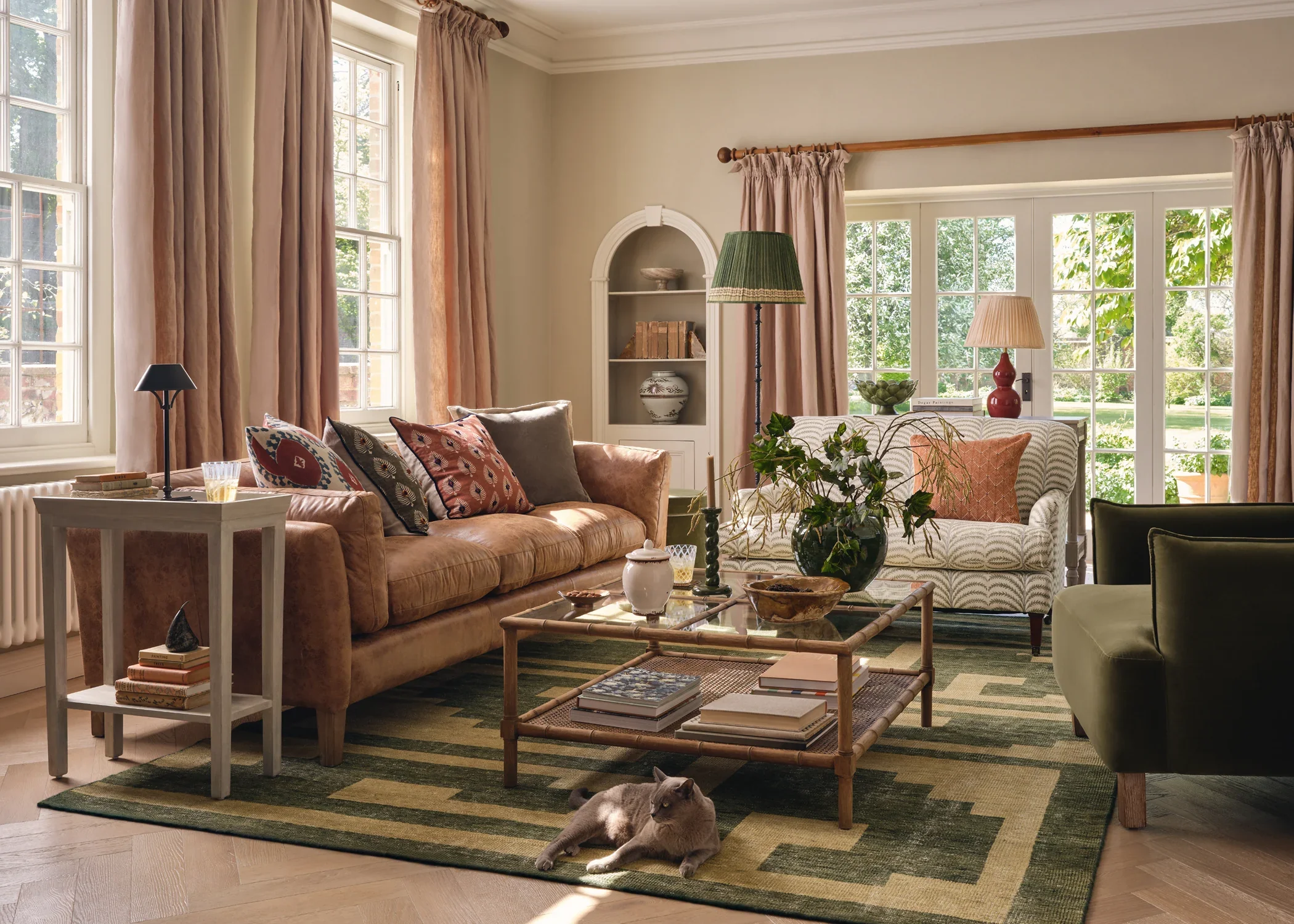 Bright living room with large windows, pink curtains, beige walls, and a mix of OKA furniture including a tan sofa and patterned loveseat. Decor includes table lamps, plants, stacked books, and a cat lying on a green-patterned rug.