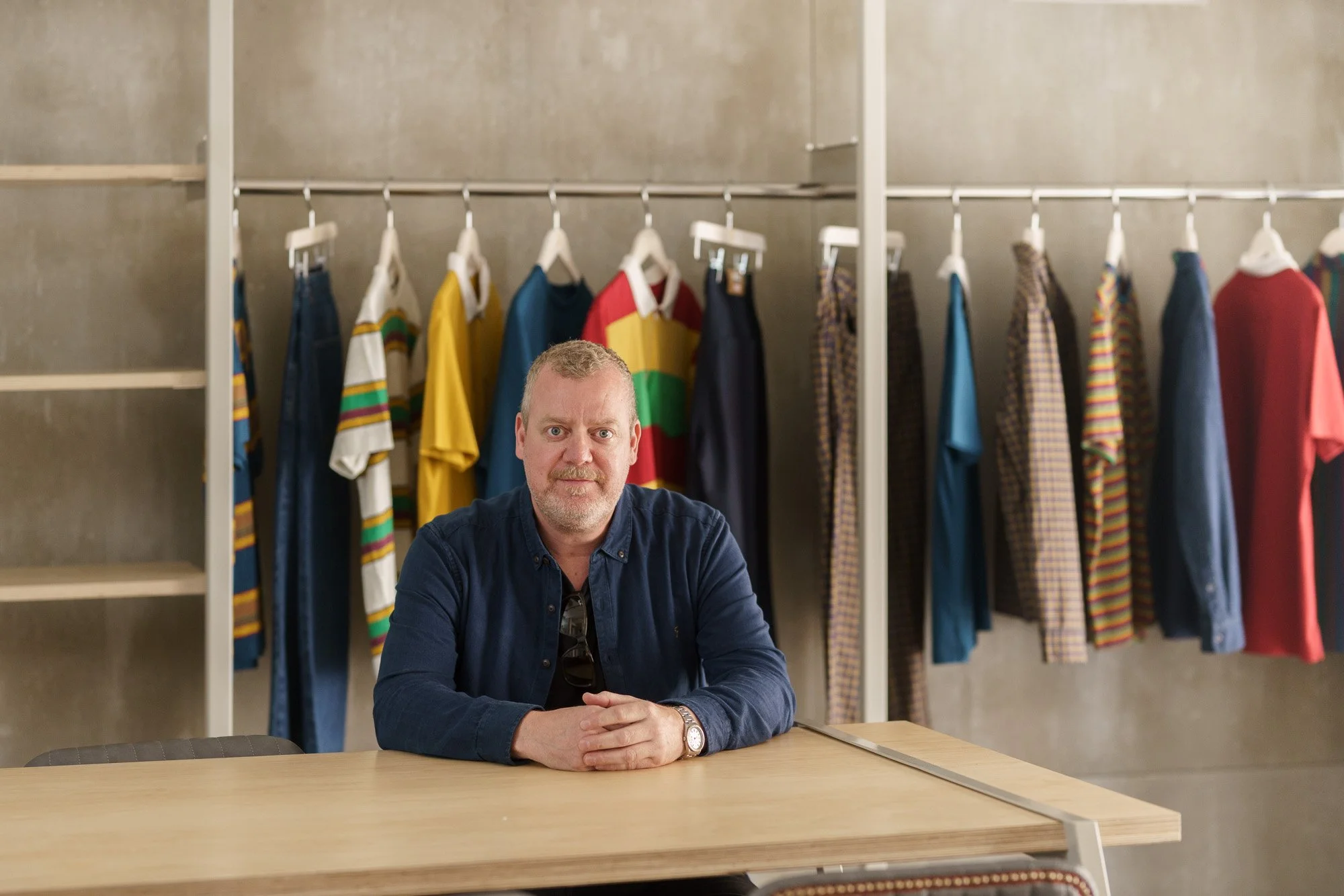 A man sitting at a wooden table in front of a clothing rack with colorful shirts and jackets.