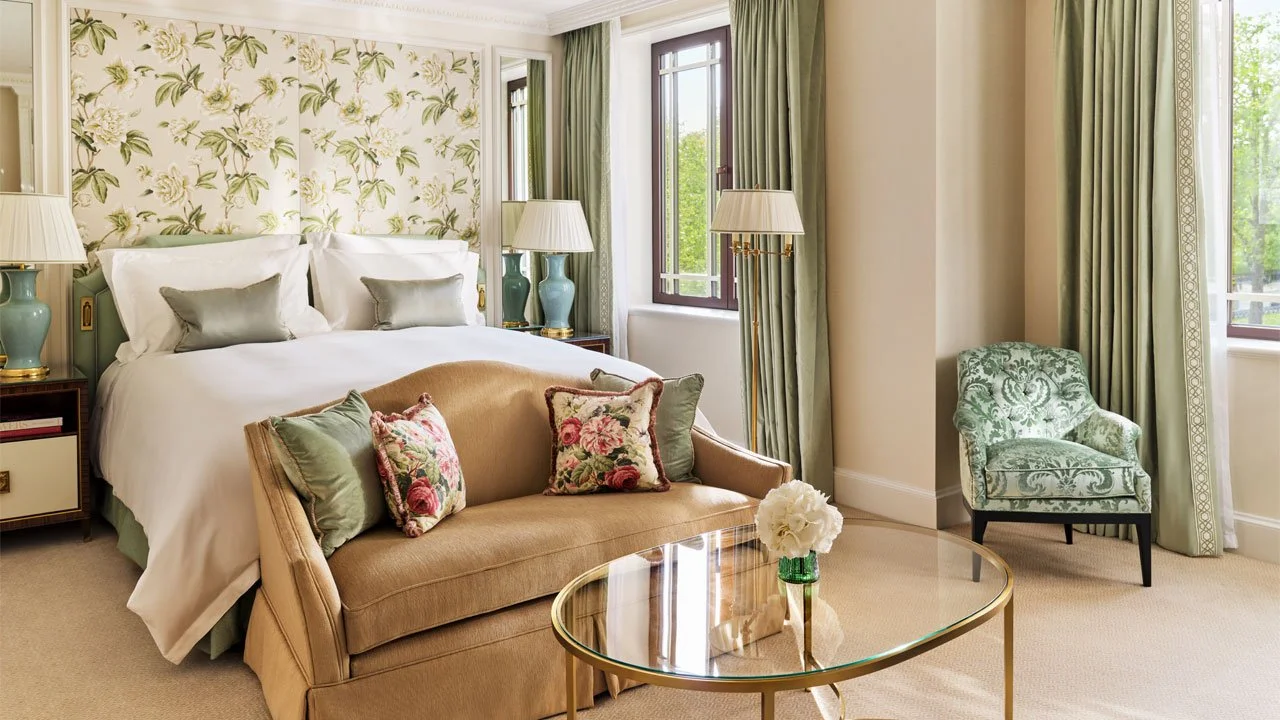 A beautifully decorated hotel bedroom with a large bed, floral wallpaper behind the headboard, green curtains, a small sofa with floral and plain pillows, a glass-top coffee table with a flower vase, and a green patterned armchair next to the window.