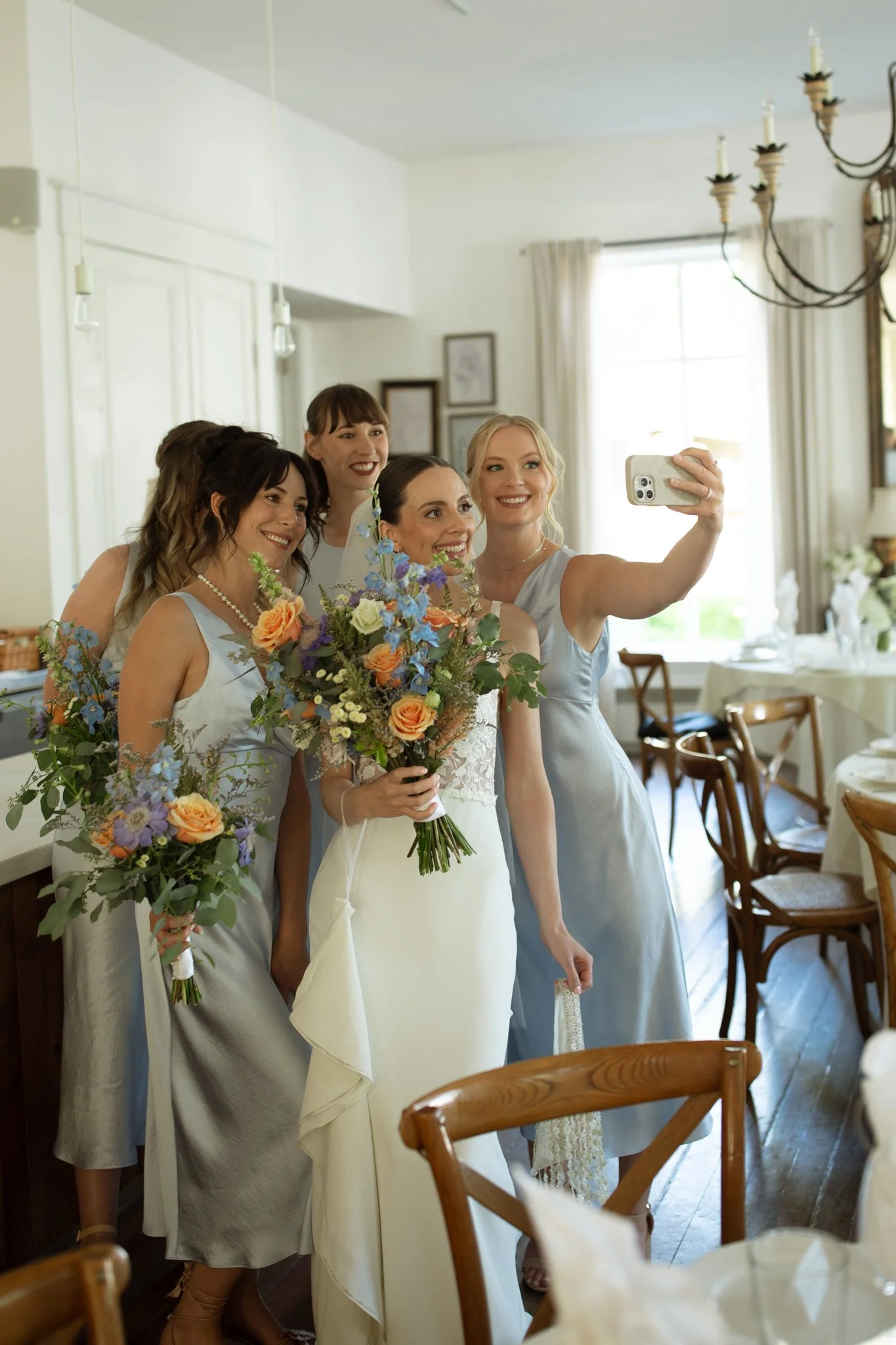 A bride taking a selfie with her bridesmaids, all holding bouquets, in a well-lit dining room with decorated tables and wooden chairs.
