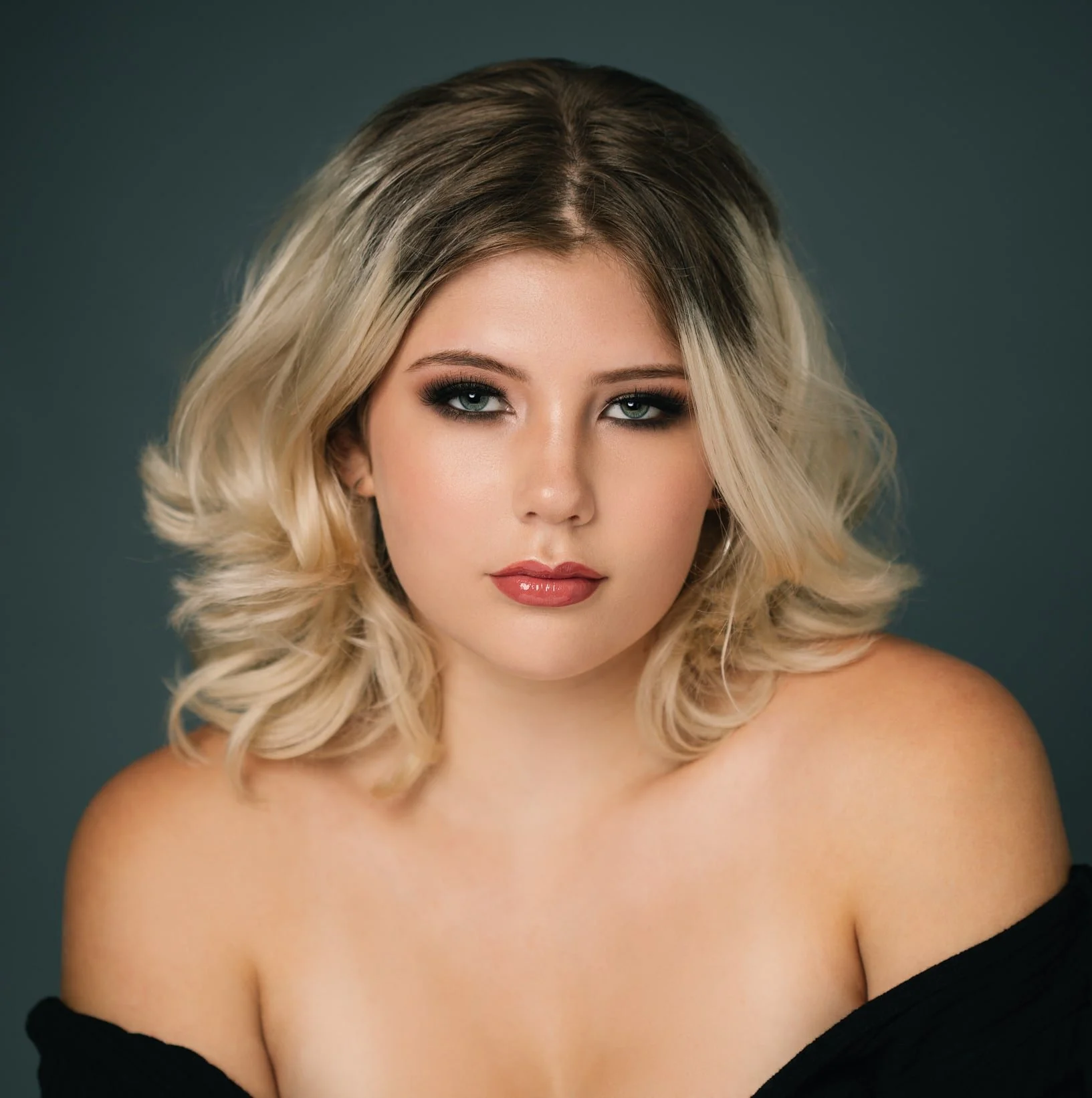 A woman with blonde, wavy hair styled in a shoulder-length bob, wearing makeup with dark eye makeup and natural lip color, looking directly at the camera against a dark background.