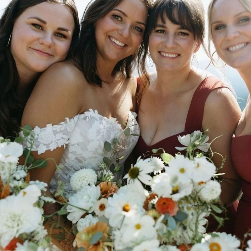 Four smiling women in formal dresses holding bouquets of flowers at a wedding or special event.