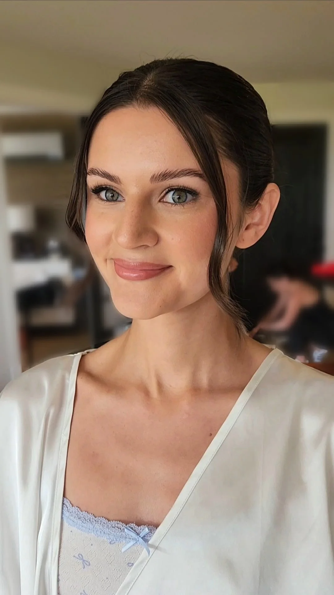 A young woman with blue eyes and dark brown hair in a loose updo, wearing a white satin robe and lace camisole, smiling softly in a well-lit room.