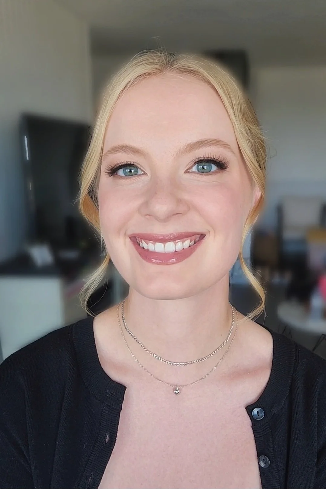 A smiling woman with blonde hair, blue eyes, wearing a black top and layered silver necklaces, in an indoor setting with blurred background.