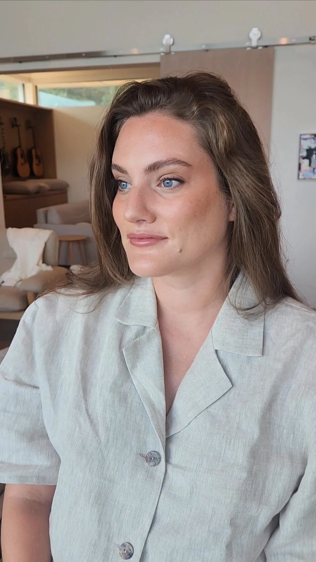 A young woman with light skin and brown hair, wearing a light beige button-up shirt, looking to her left in an indoor setting with guitars on the wall and a couch in the background.