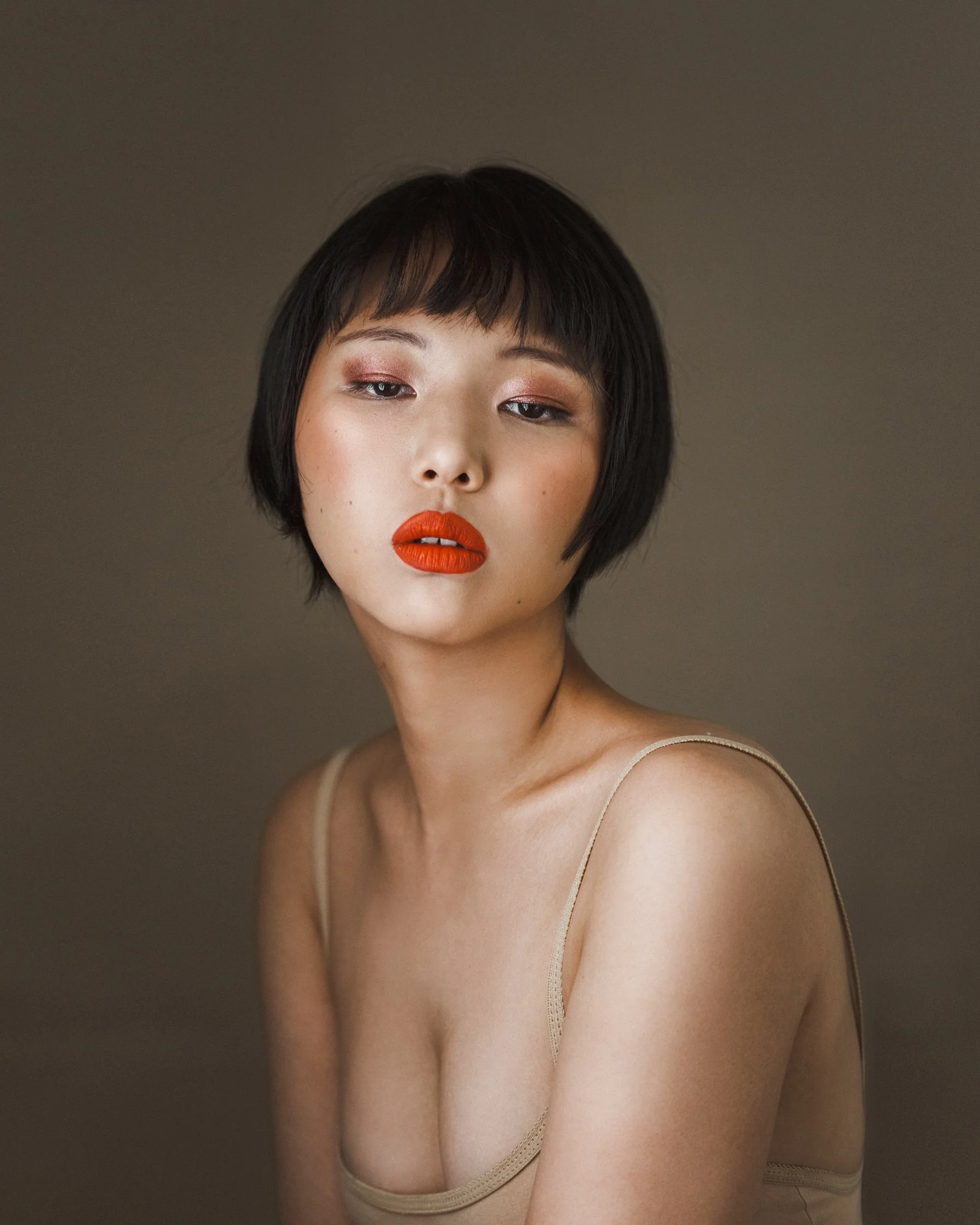 A woman with short black hair and red lipstick, wearing a beige spaghetti strap top, posing against a neutral background.