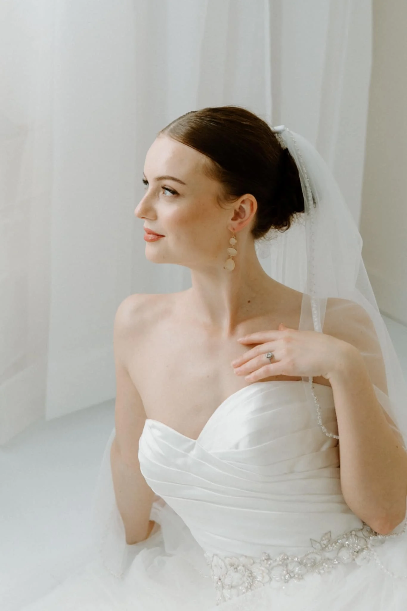 A bride in a strapless white wedding dress with satin and beaded embellishments, wearing pearl drop earrings, a wedding ring, and a white veil with a black bow, looking to the side, in a softly lit room with white curtains.