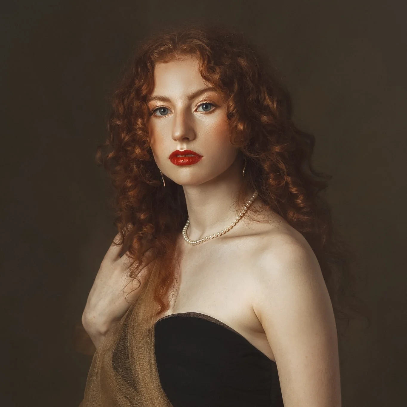 A woman with red, curly hair wearing red lipstick, pearl necklace, and earrings, dressed in a strapless black and gold outfit, against a dark background.