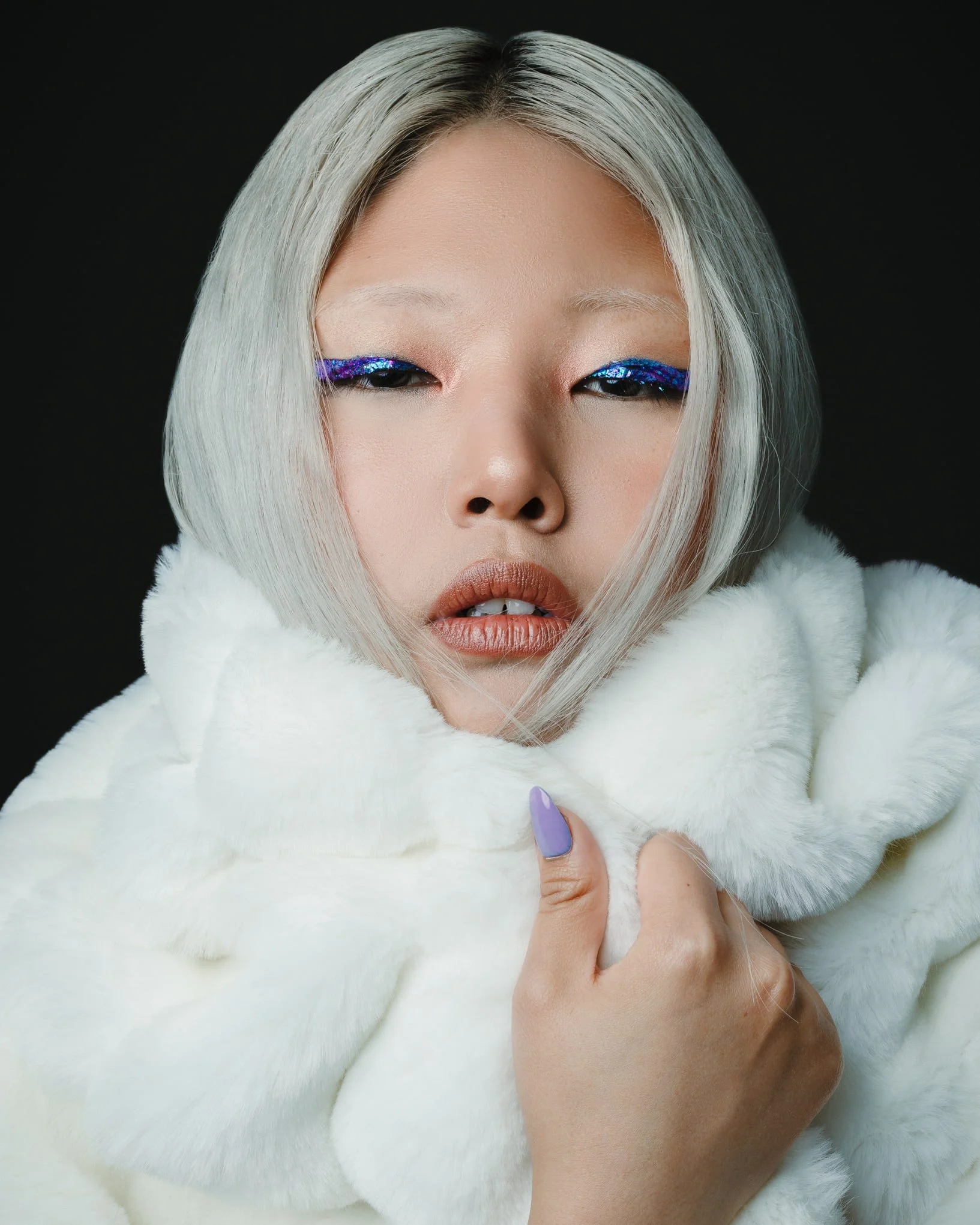 Close-up of a woman with platinum blonde hair, glossy lips, and vibrant purple and blue eye makeup, wrapped in a white faux fur garment against a black background.