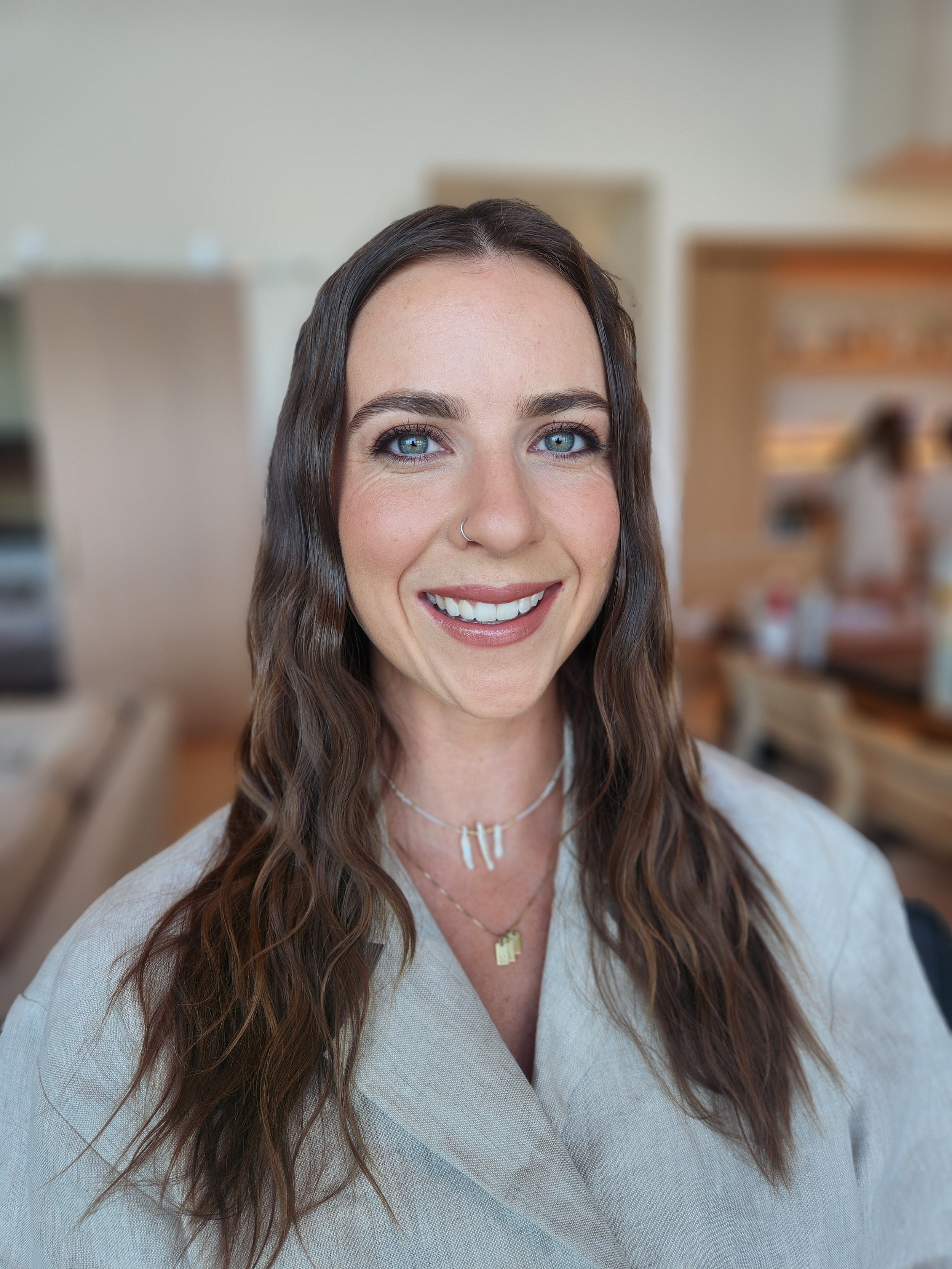 A woman with long brown wavy hair, blue eyes, and a nose piercing, smiling indoors with blurred background.