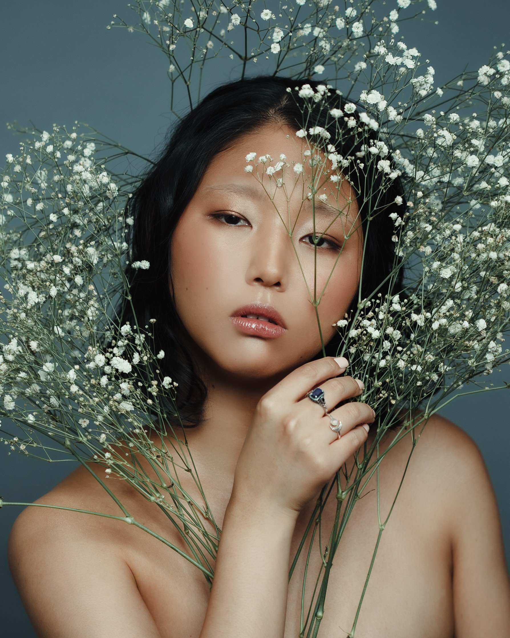 A woman with black hair and fair skin holding white flowers around her face, with a serious expression, soft makeup, and rings on her fingers against a dark background.
