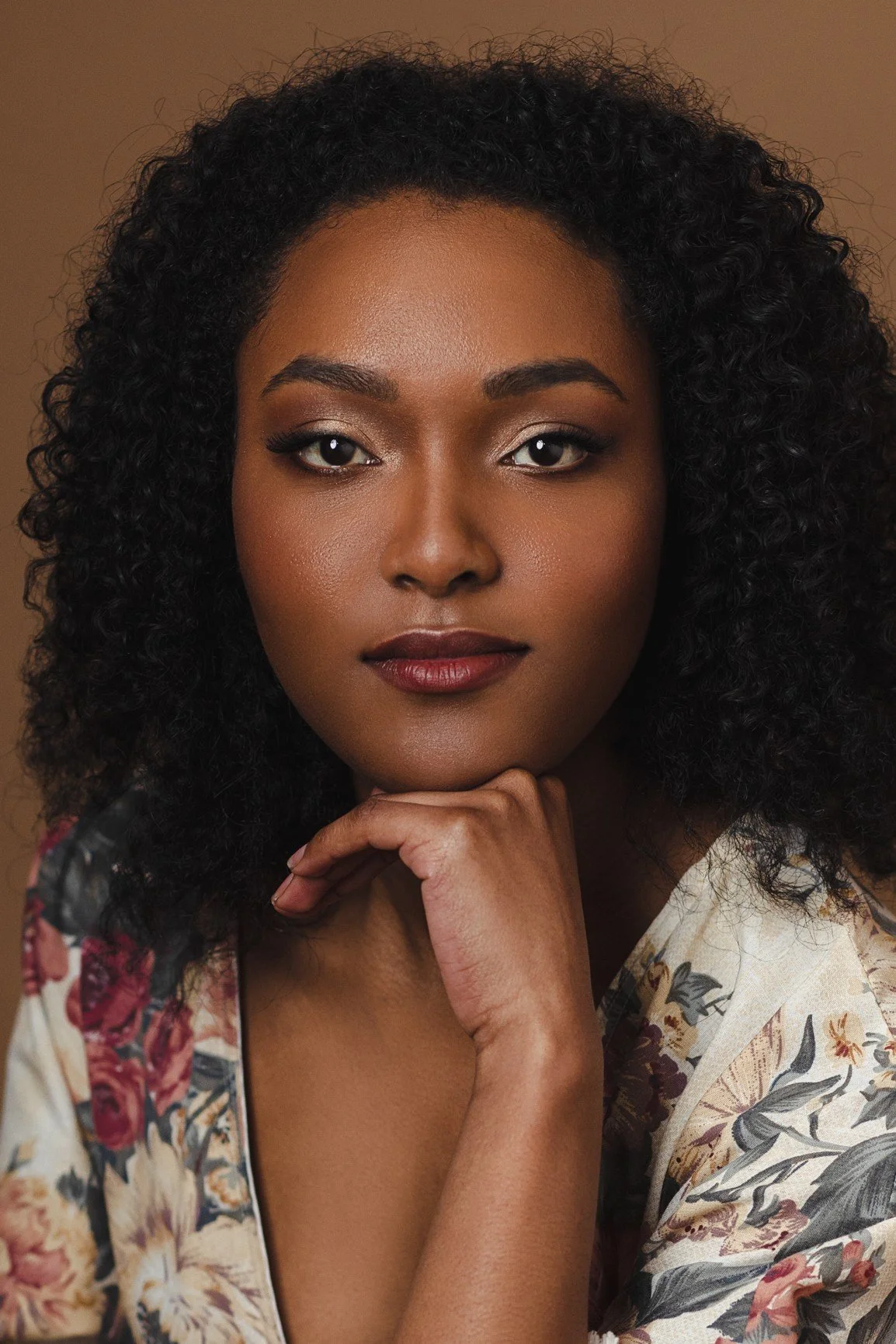 A close-up portrait of a woman with curly black hair, wearing makeup, and a floral-patterned top, with her hand resting under her chin.