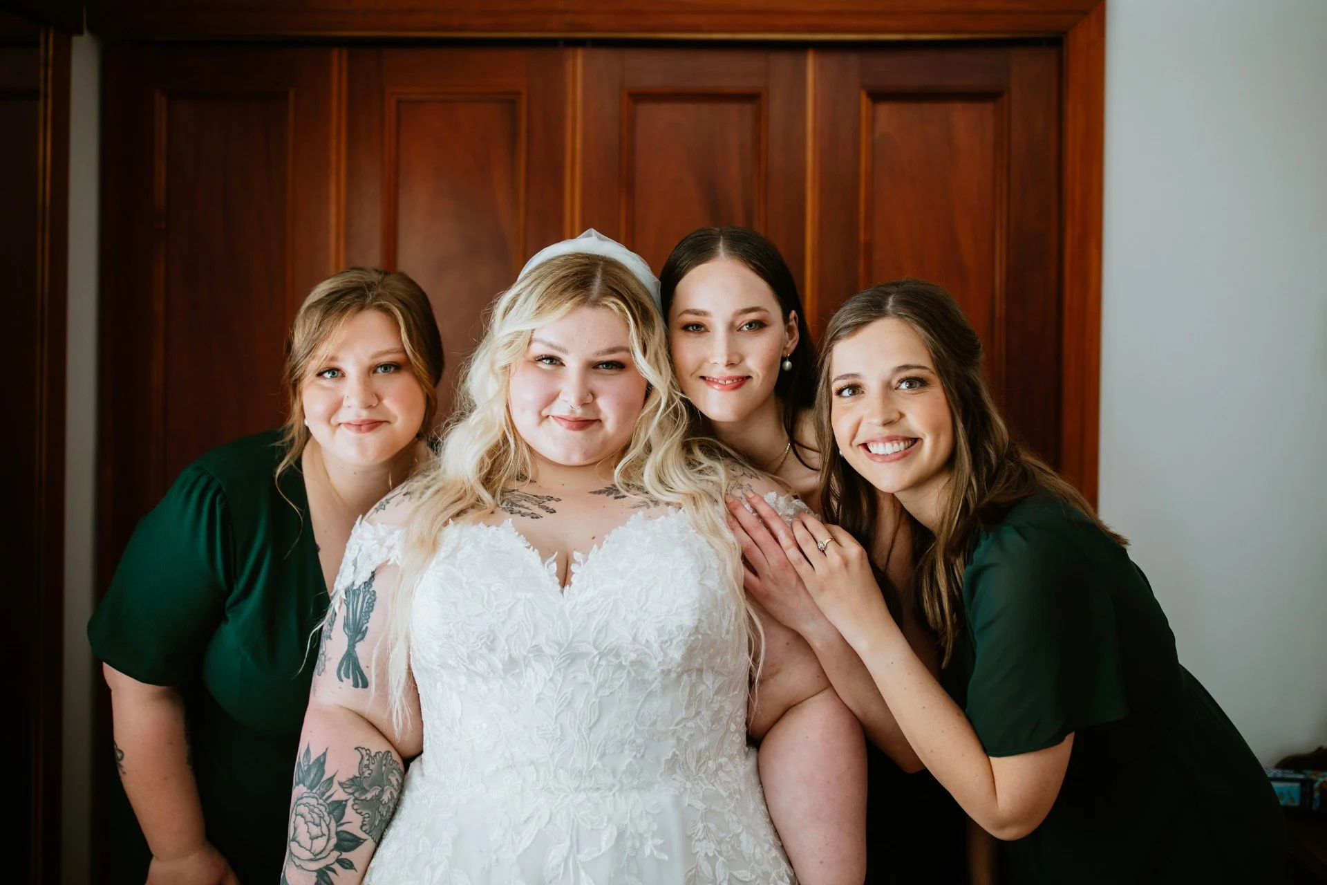 Four women smiling, one in a wedding dress, just before her wedding ceremony, standing in front of a wooden door.