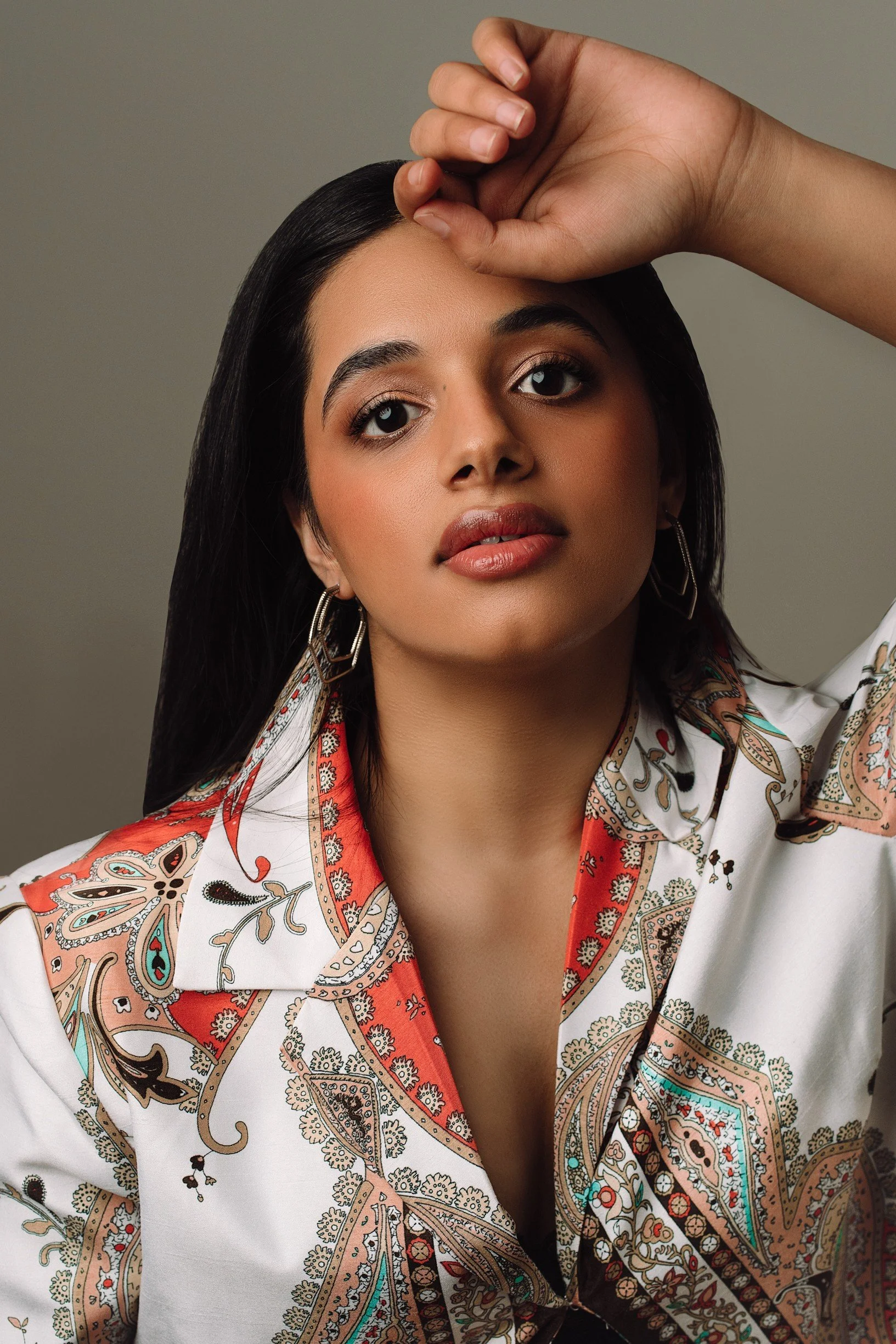 A woman with long dark hair, wearing a patterned shirt with paisley and floral designs, posing with her hand resting on her head against a plain background.