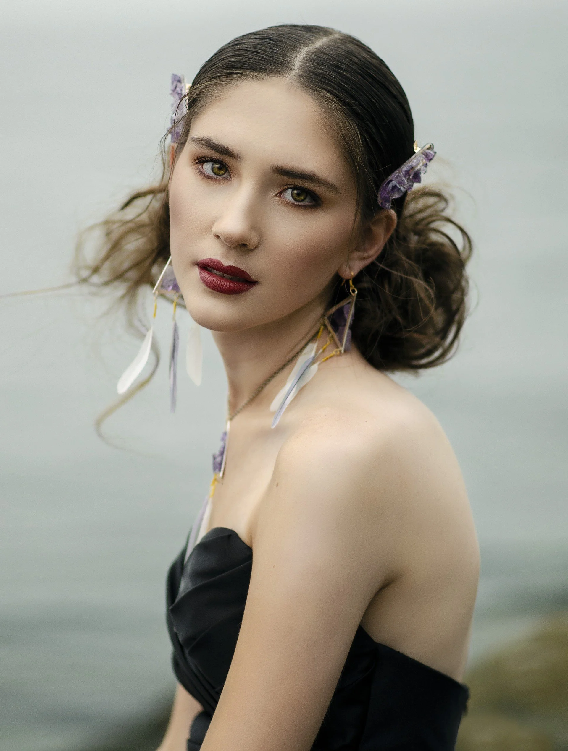 A young woman with dark hair, wearing purple hair clips, earrings, and a black strapless dress, looks at the camera near water with a blurred background.