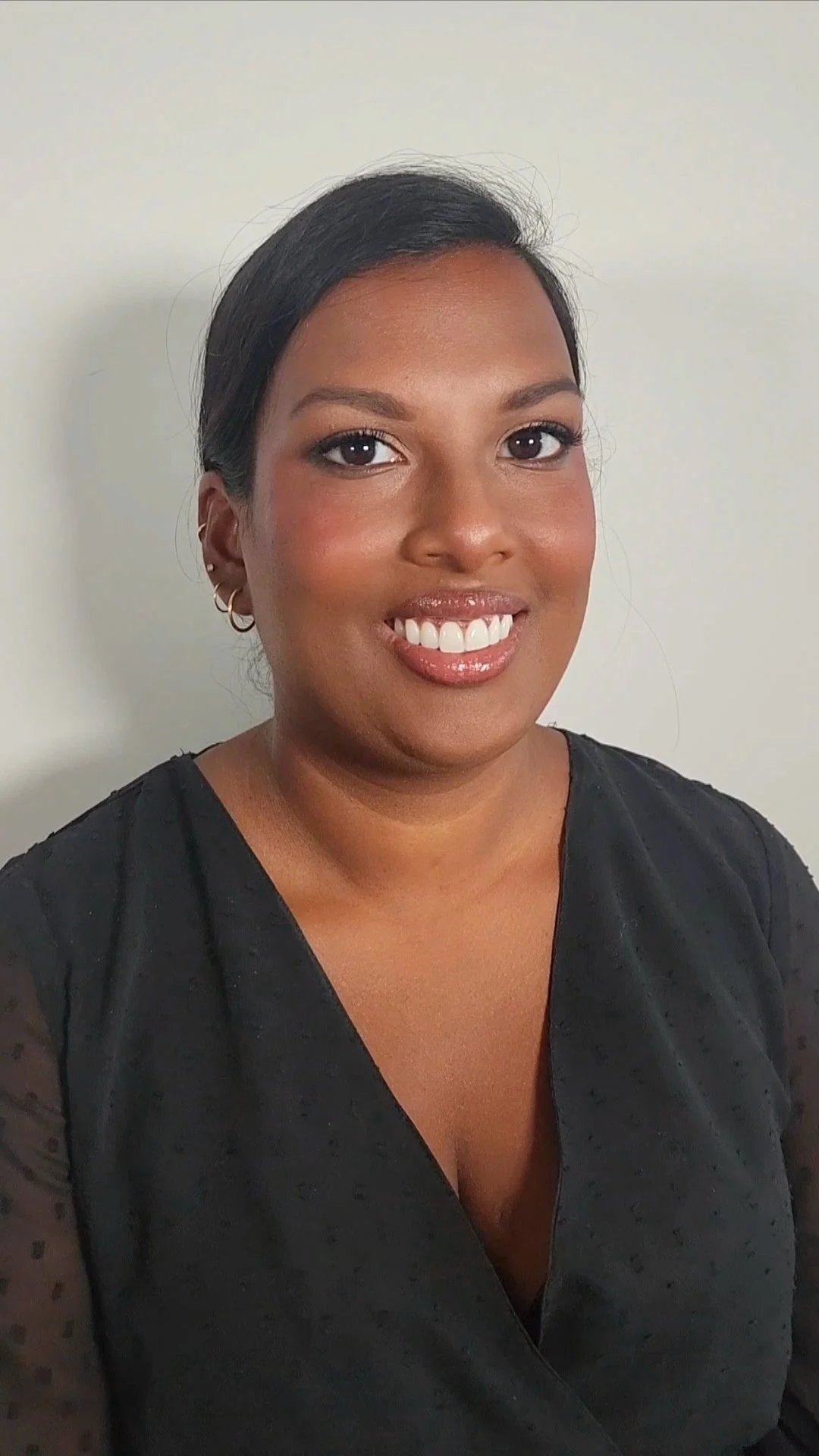 A woman with short dark hair, brown eyes, and medium skin tone smiling in front of a plain white wall, wearing hoop earrings and a black top.
