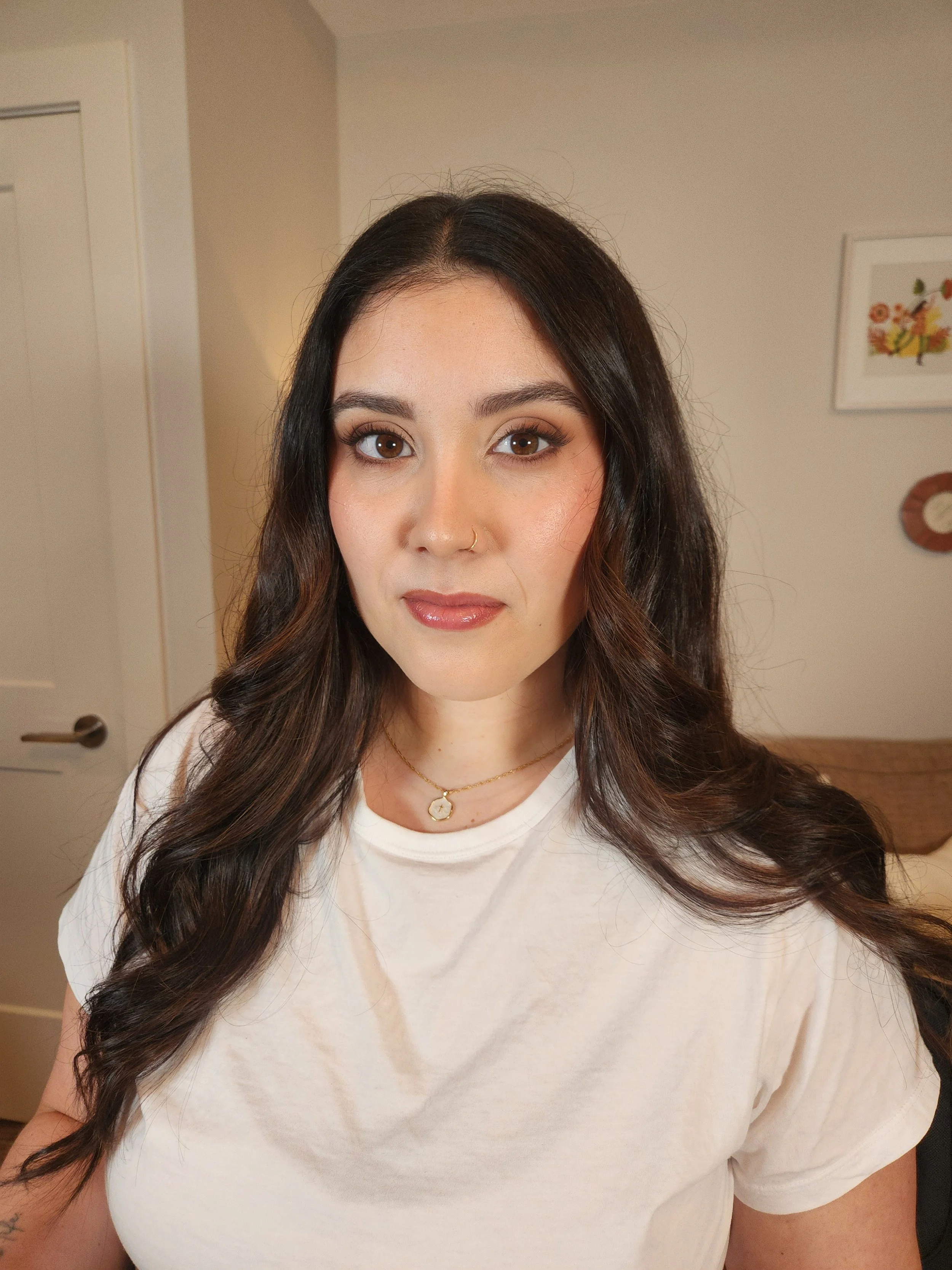 A woman with long, dark wavy hair and a nose piercing, wearing a white t-shirt and gold jewelry, standing in a home interior.