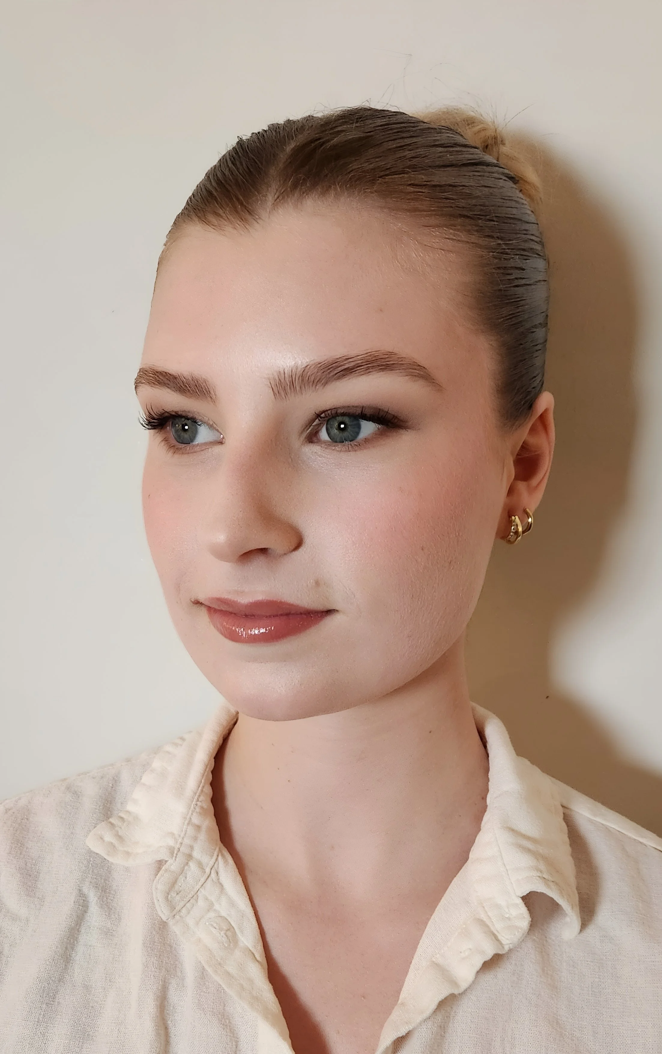 A close-up portrait of a young woman with light skin, blonde hair tied back, blue eyes, wearing a beige shirt and gold earrings, standing against a plain white background.