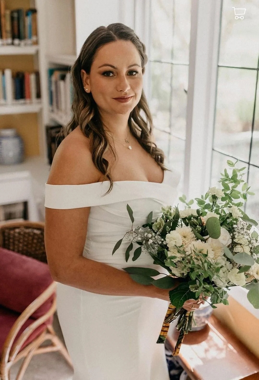 A woman in a white off-the-shoulder dress holding a bouquet of white flowers, standing indoors near large windows.