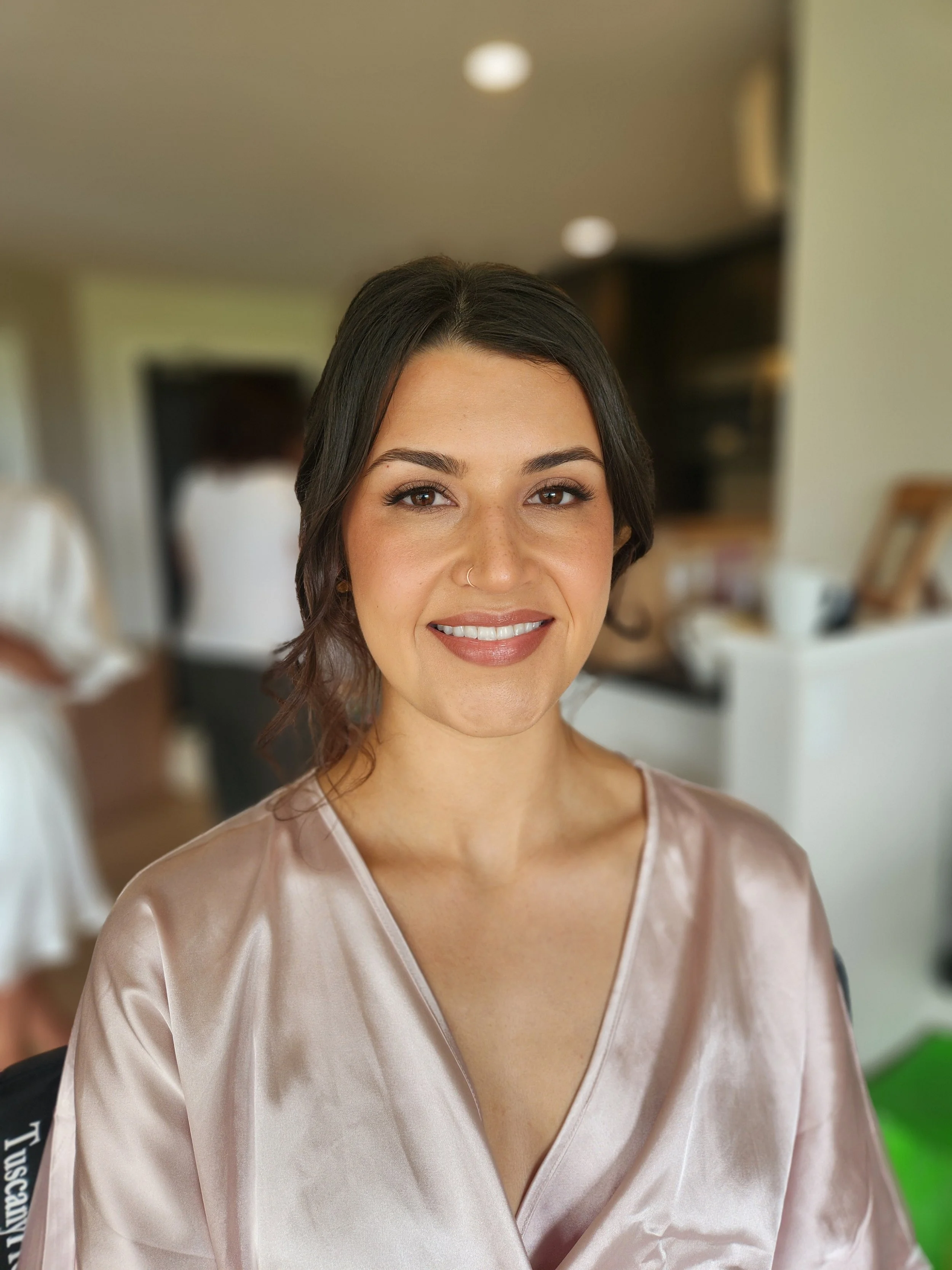 A young woman with dark hair, makeup, and a nose ring, smiling at the camera inside a room with blurred background.
