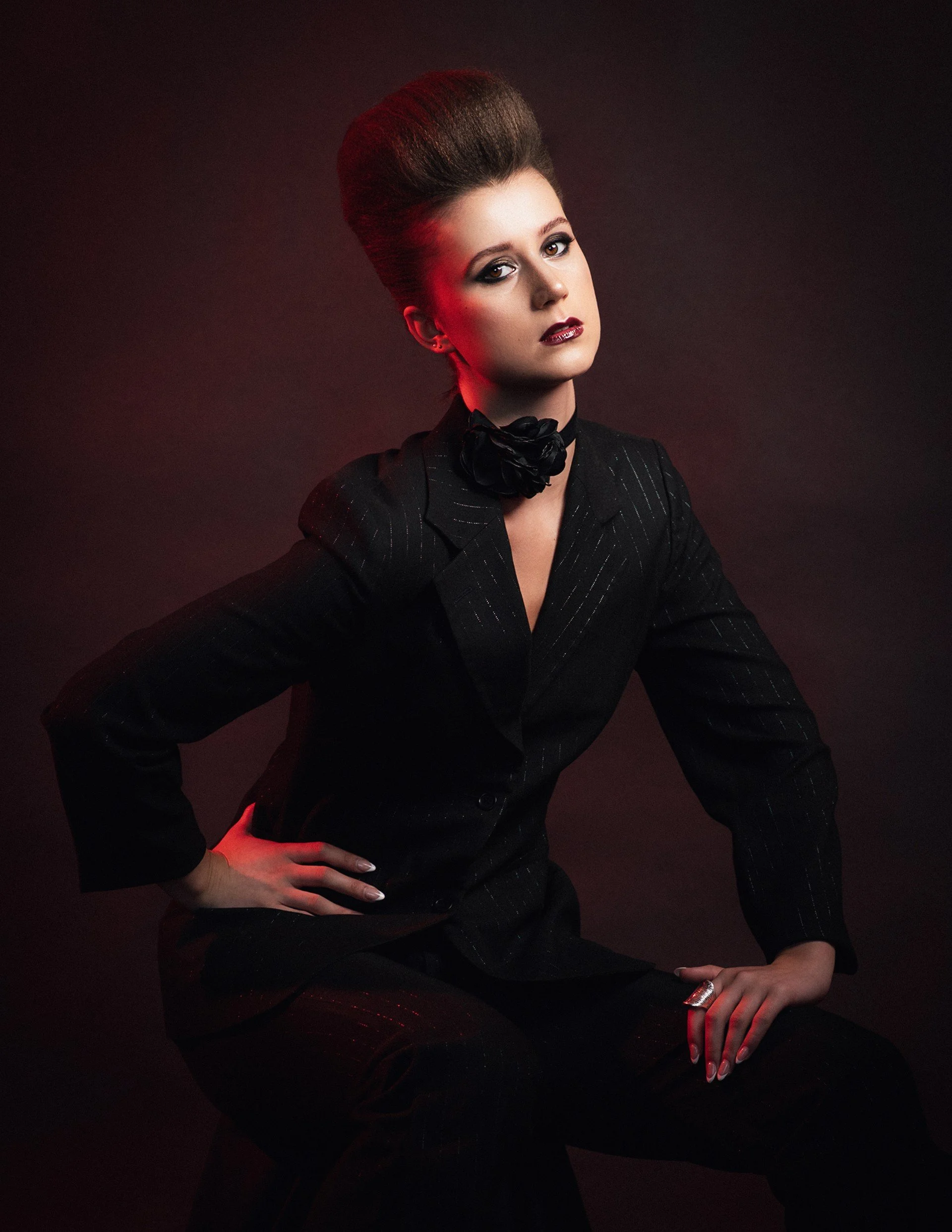 A woman with styled hair wearing a black pinstripe suit and a black flower neck accessory, sitting against a dark background with red lighting.