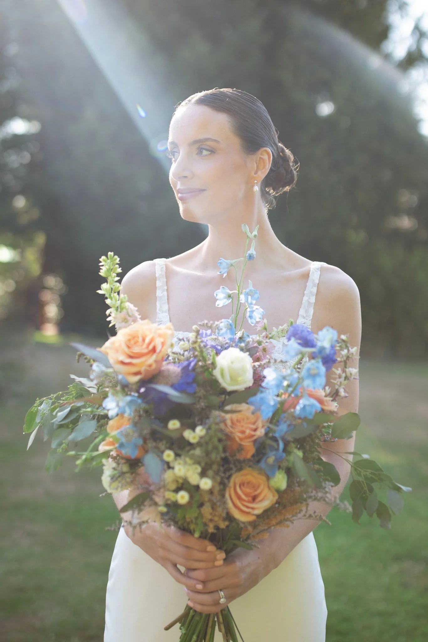 A woman in a white dress holding a large bouquet of colorful flowers outdoors, with sunlight streaming in from the upper left corner.