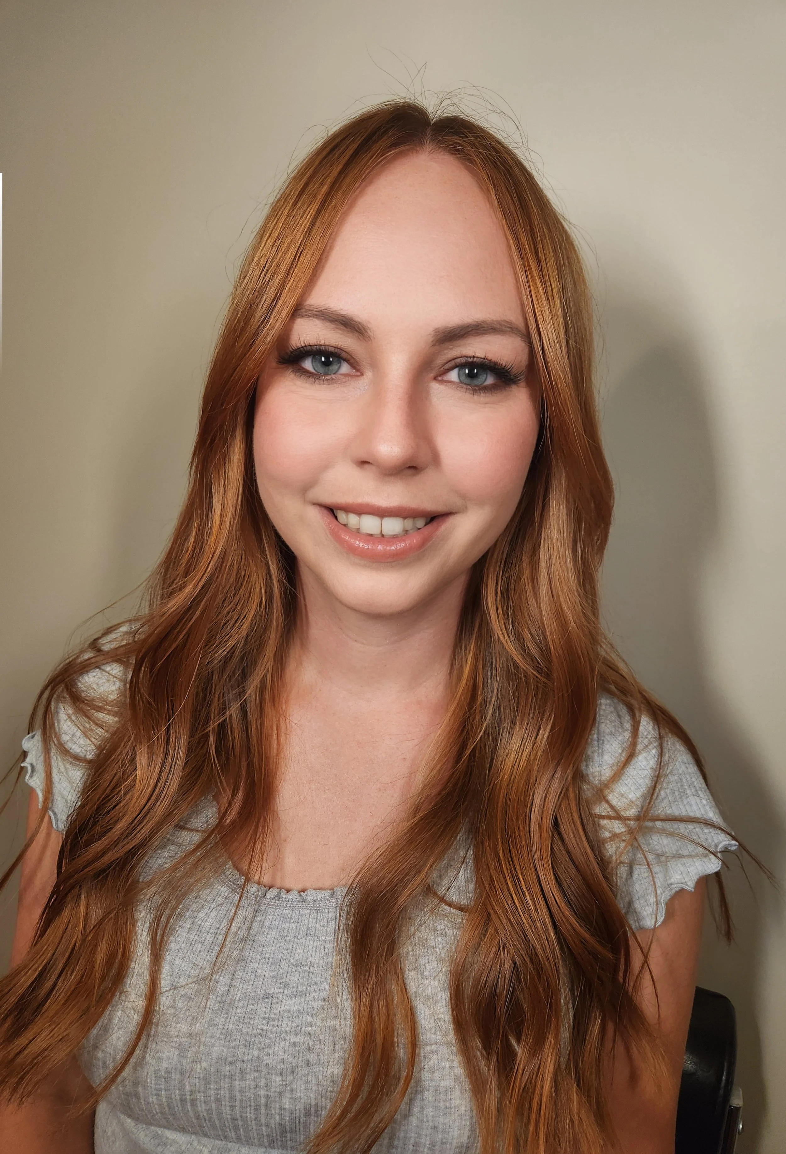 Close-up of a young woman with long, wavy red hair, blue eyes, and light makeup, smiling gently at the camera against a plain beige wall.