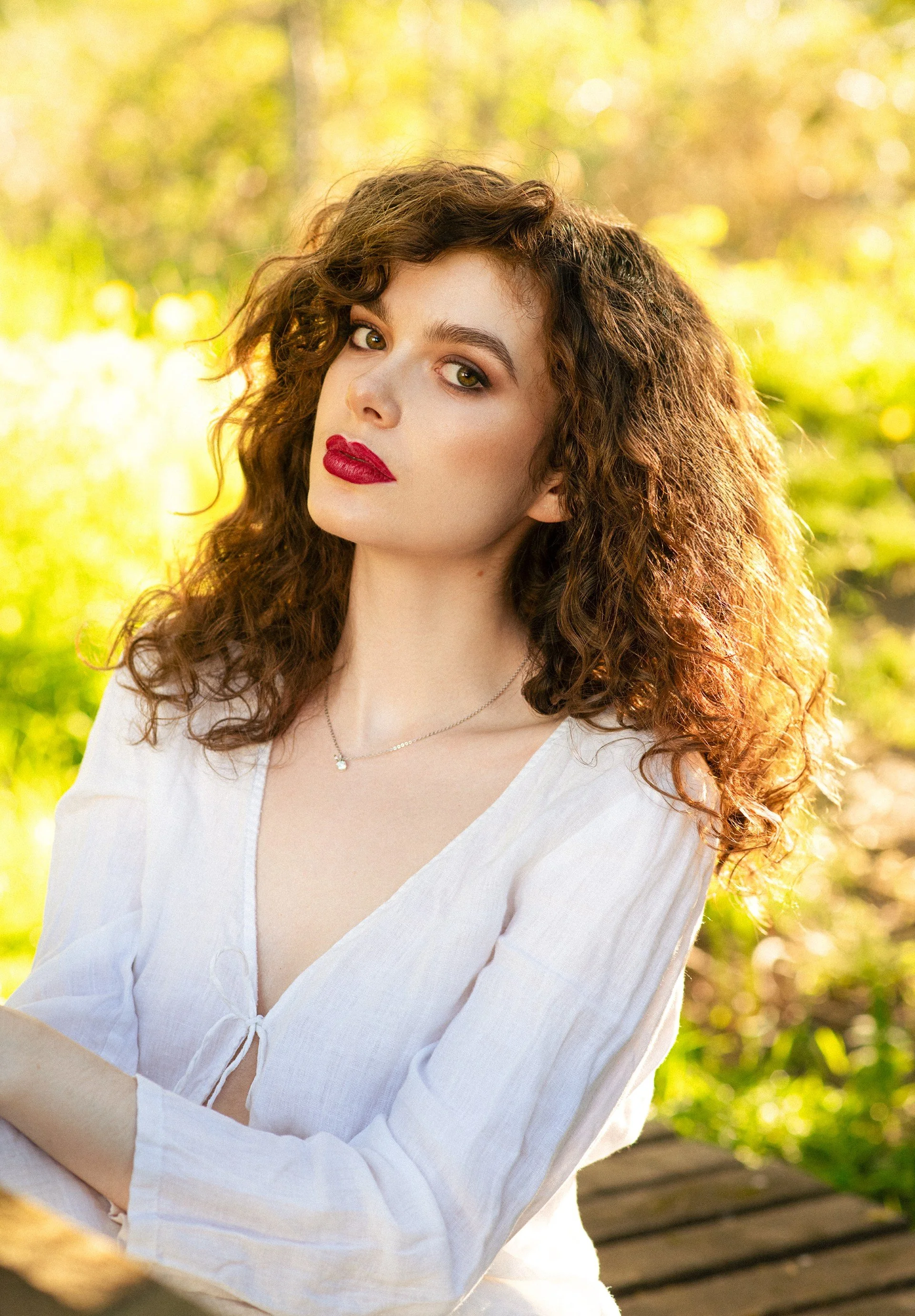 A woman with curly brown hair and red lipstick is sitting outdoors on a wooden bench, wearing a white blouse, with a blurred natural background of yellow and green foliage.