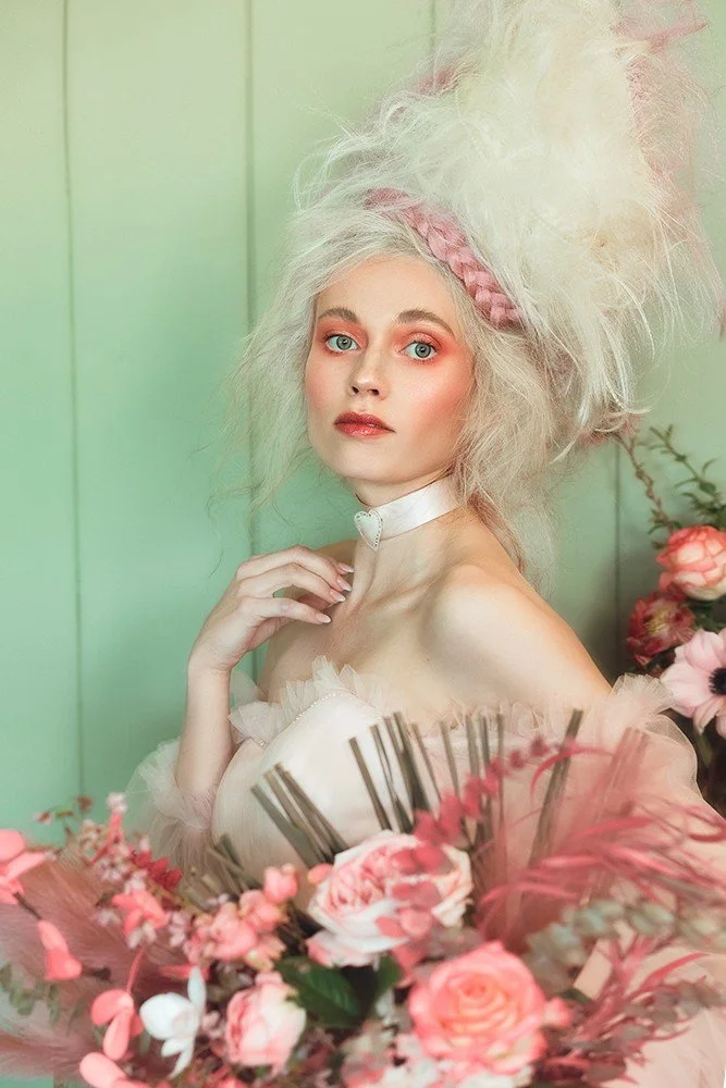 A young woman with platinum blonde, voluminous hair styled in an elaborate updo with pink accents. She has fair skin, light makeup with pink blush and lip color, built-in eyebrows, and striking blue eyes. She is wearing a white choker with a heart-sh