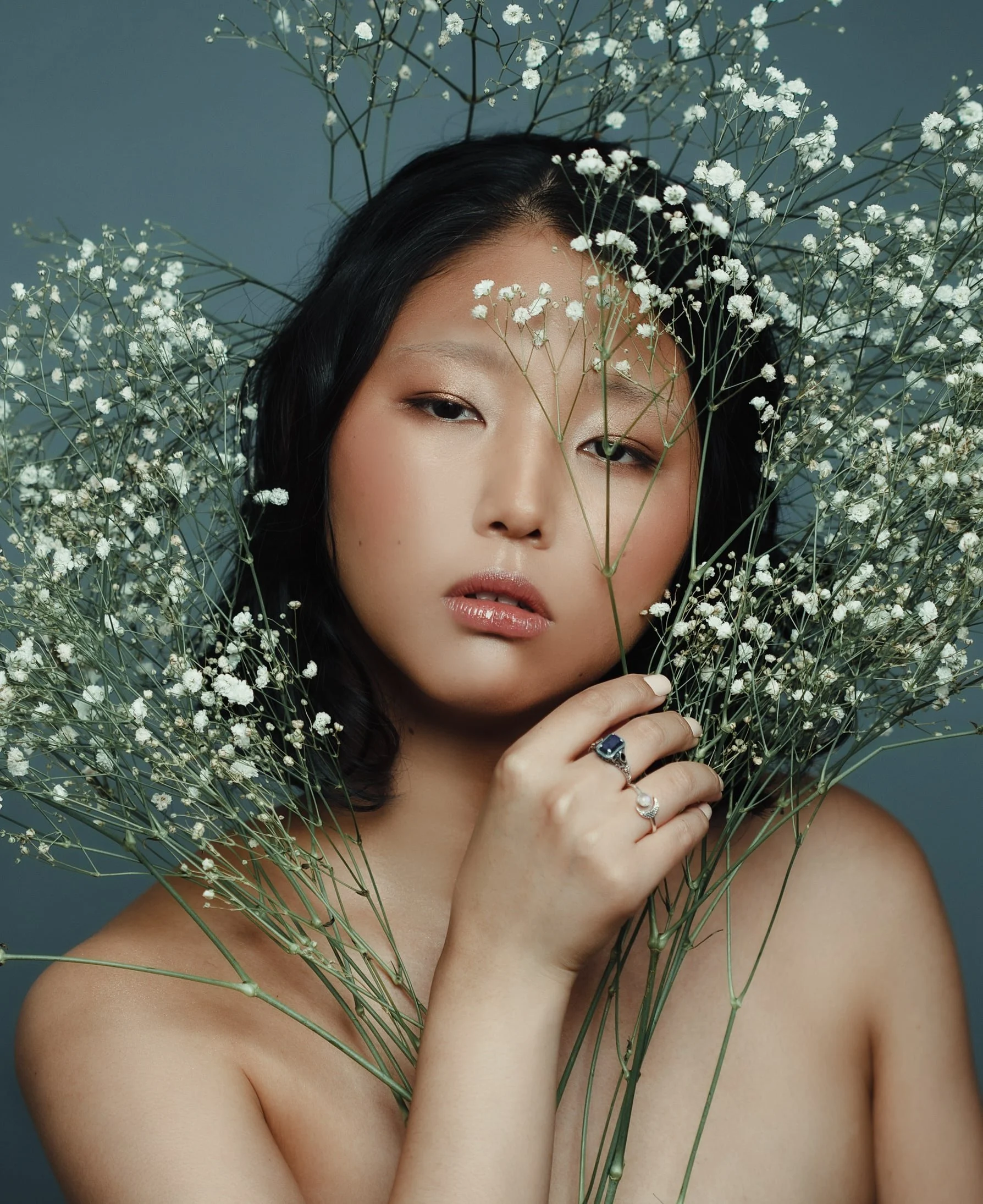 A woman with dark hair holding a stem of white flowers, surrounded by more flowers, against a muted blue background.