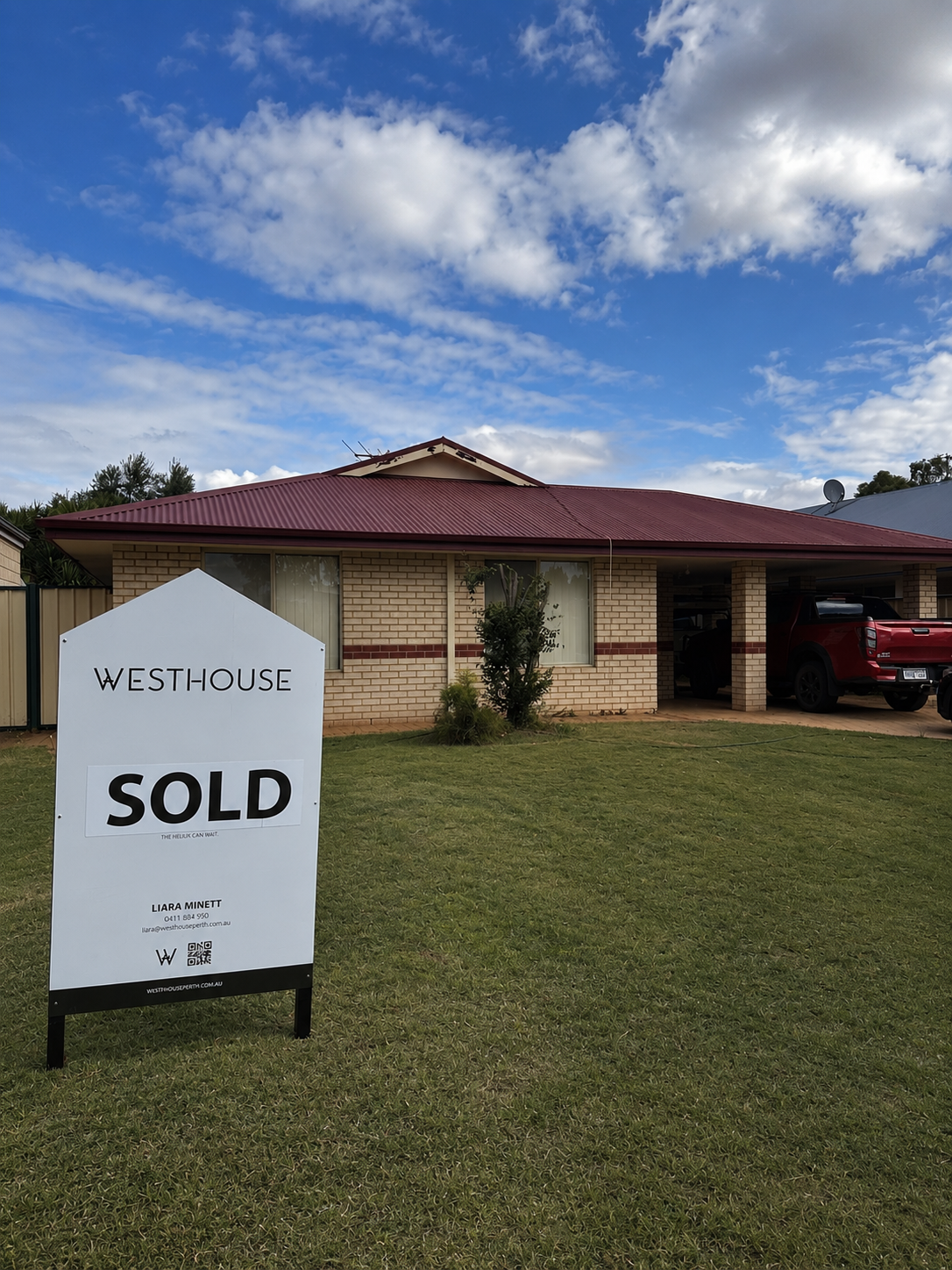 A brick house with a red roof and large windows, parked red truck in the garage, small tree in front, and a sold sign on the lawn indicating the house has been sold.